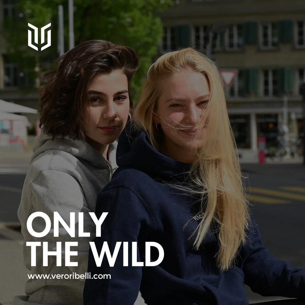 Two young women outdoors, one with brown hair and the other with blonde hair, smiling at the camera. They are dressed casually, with a city street and buildings in the background. A logo and the text 'ONLY THE WILD' and 'www.veroribelli.com' are over