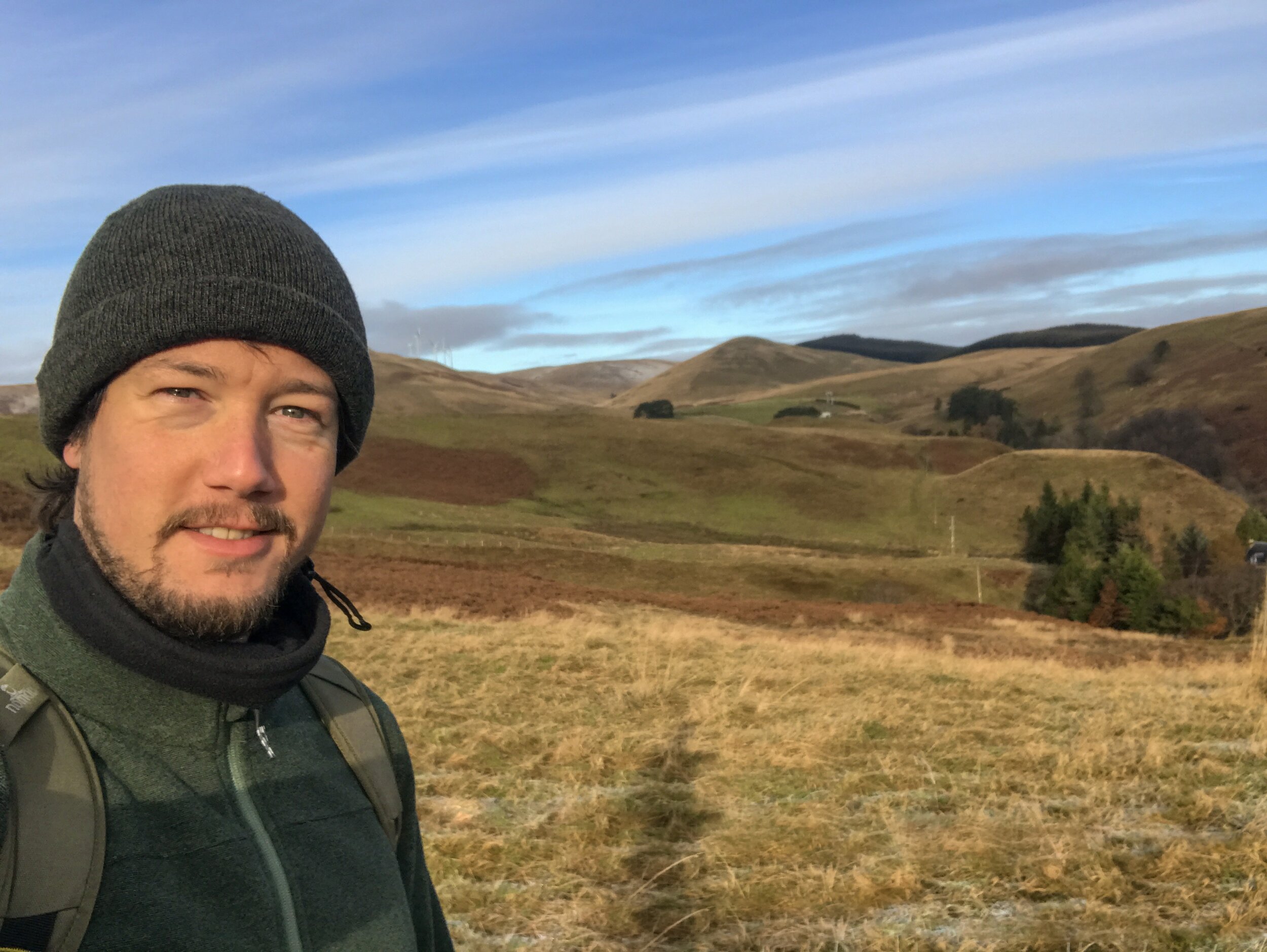A man wearing a dark beanie and black jacket, smiling, standing in a grassy, hilly landscape with patches of trees and a blue sky with clouds in the background.