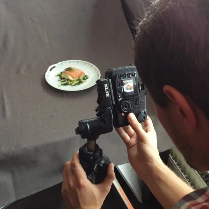 Person taking a photograph of a gourmet dish on a white plate, which contains a piece of seafood and green garnish, using a professional camera.