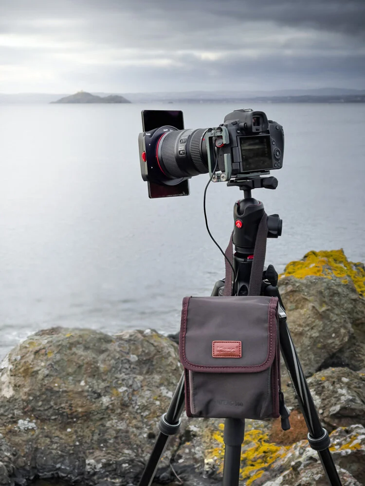 Camera on a tripod set up on rocky shore overlooking a body of water with an island in the distance under a cloudy sky.