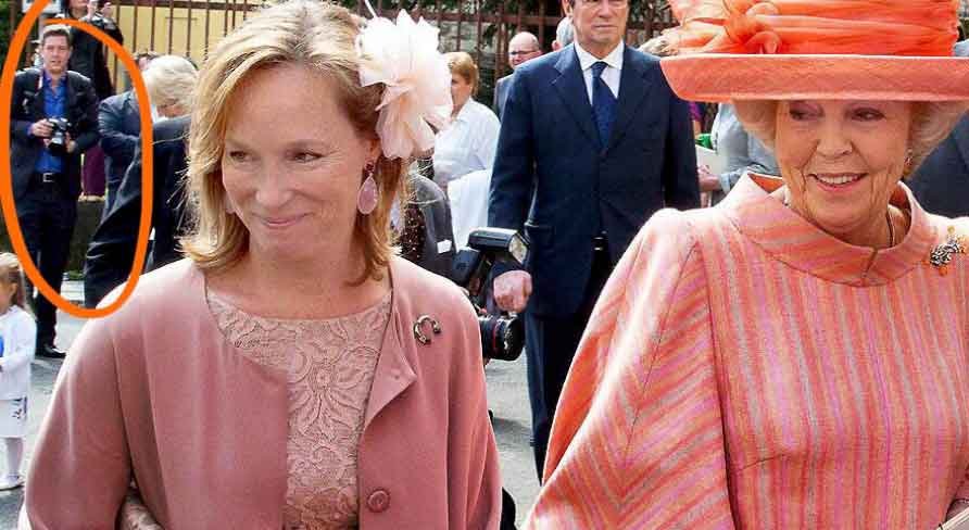 A woman with blonde hair, wearing a pink dress and jacket with a large pink flower in her hair, smiling and looking to her left. Other people, including a woman in a large pink hat and orange dress, are in the background at a public event.