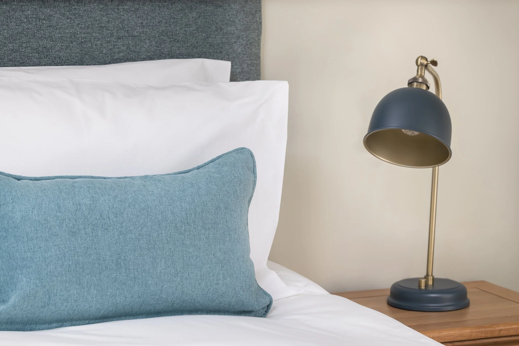 Close-up of a bed with white pillows and a blue lumbar cushion, with a wooden nightstand and a black and brass desk lamp against a cream-colored wall.
