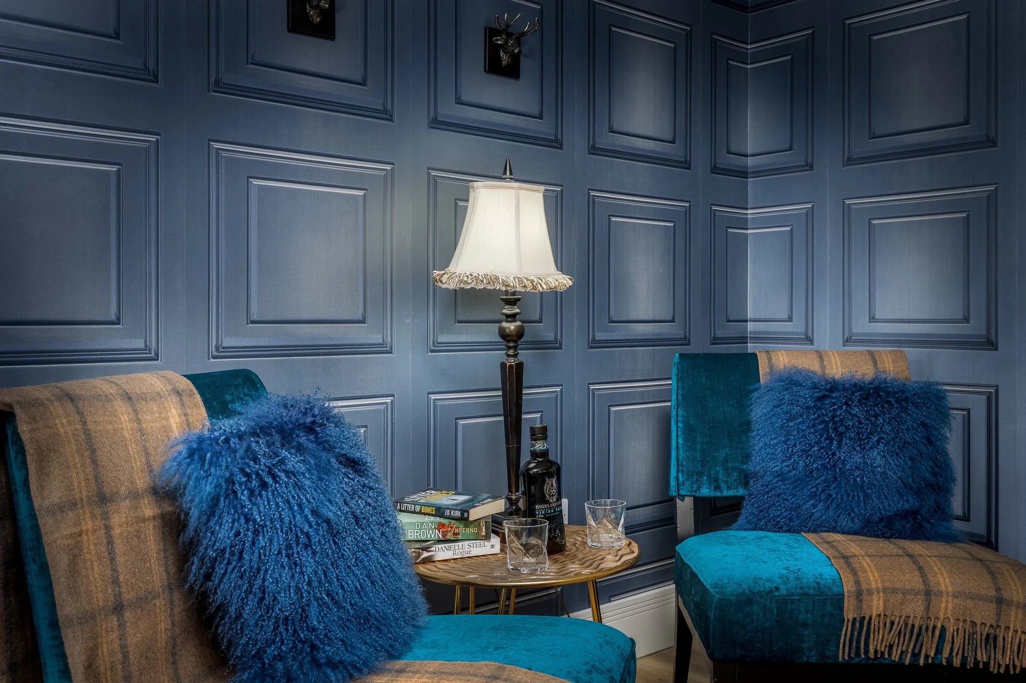 Cozy corner at Orcadia, Dornoch, a holiday rental, with two teal and tan plaid armchairs with blue pillows, wooden side table with books and glasses, vintage table lamp, and blue paneled wall with mounted decorative stag heads.