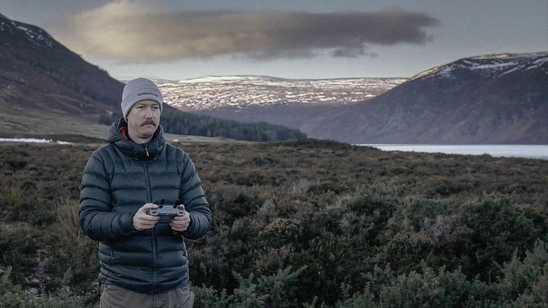 Man holding a remote controller in a mountainous landscape with snow-capped peaks and a lake in the background.