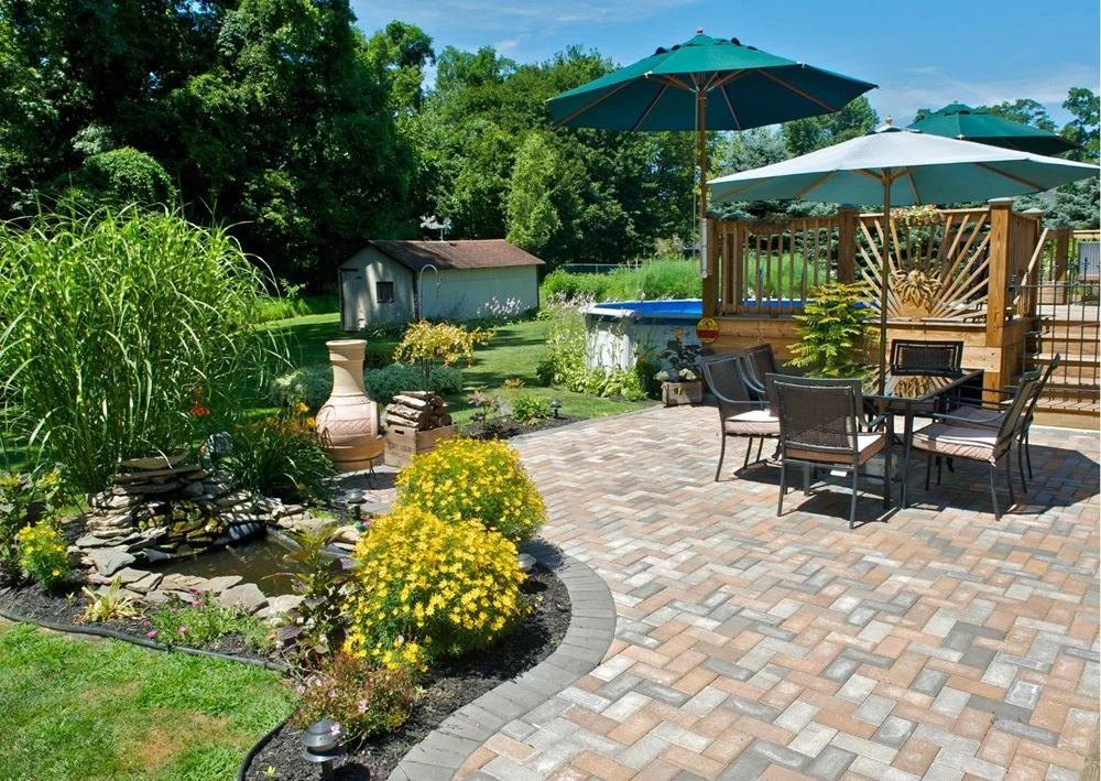 Brick patio with outdoor dining furniture, colorful umbrellas, garden beds, water feature, and sunny landscaping.