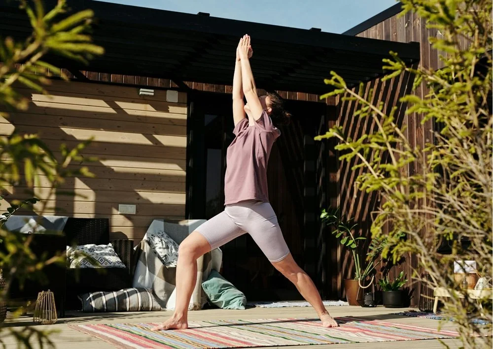 Covered patio with a person practicing yoga, outdoor cushions, colorful rug, wood walls, and surrounding plants.