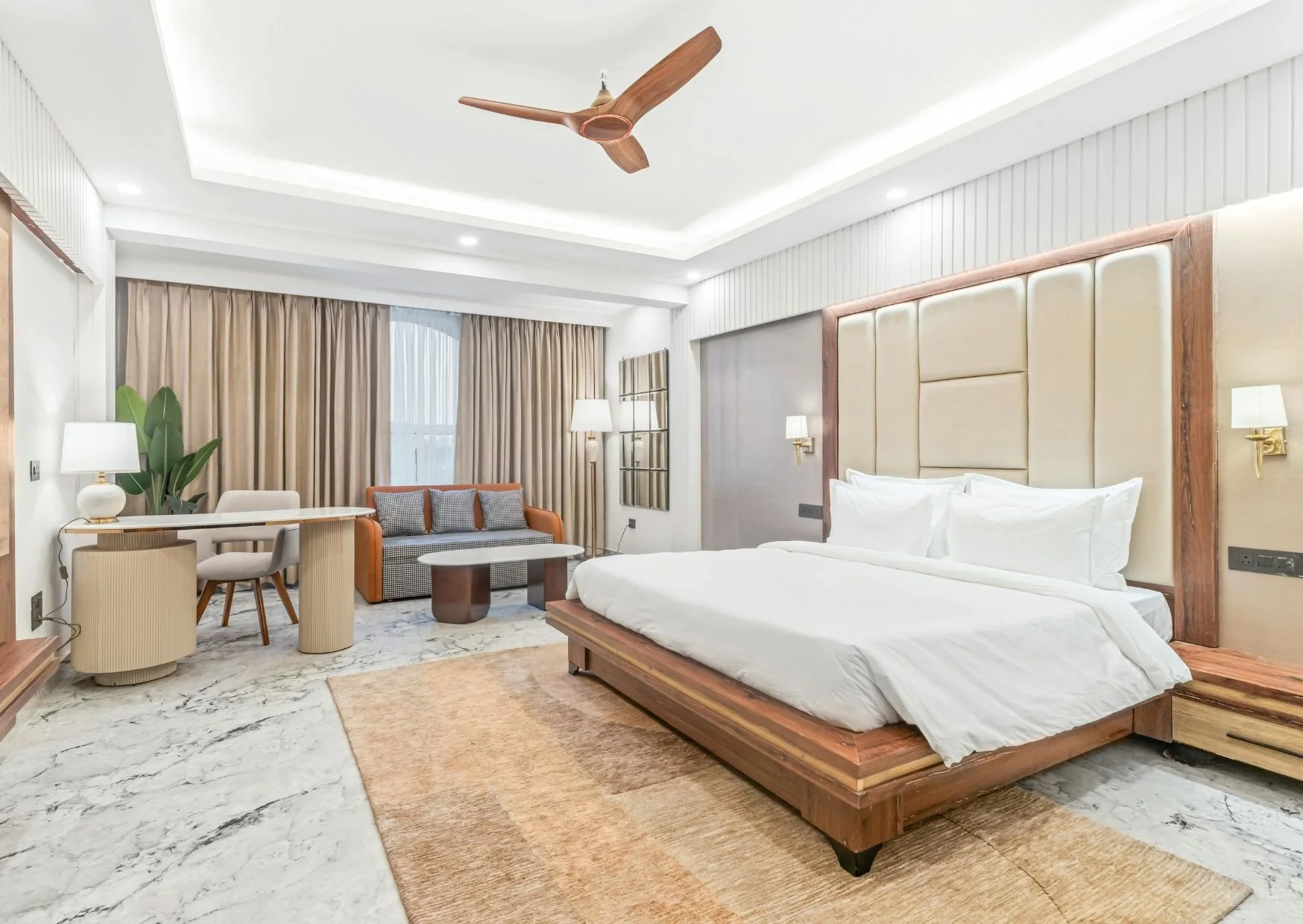 Spacious hotel-inspired bedroom with wooden platform bed, sitting area, marble flooring, ceiling fan, and elegant neutral palette.