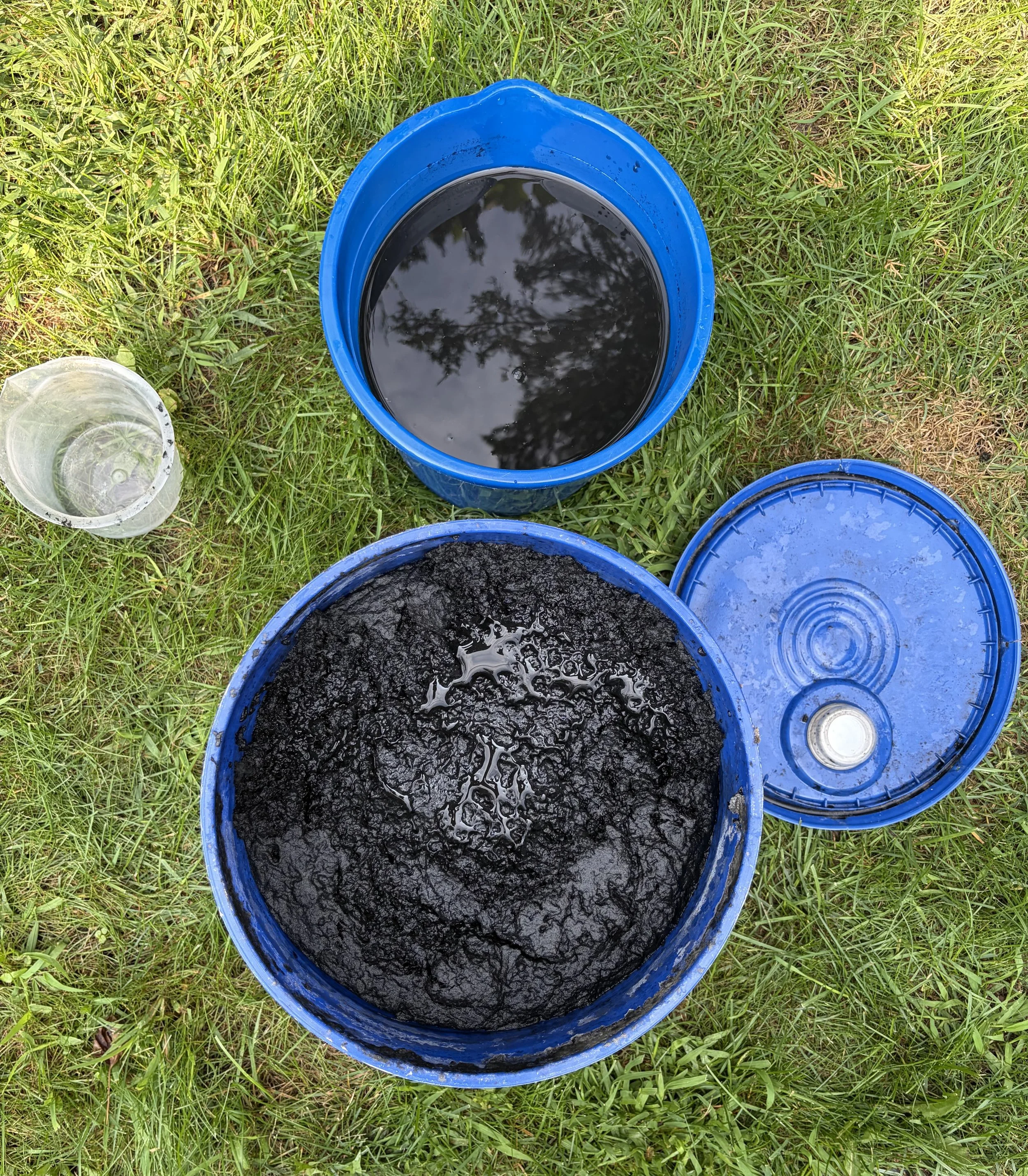 5 Gallon pail of concentrated Black Liquid Mulch for Landscaping beside a smaller pail with diluted Black Liquid Mulch for Landscaping. 1 L plastic container used to measure the Liquid Mulch and the water (5 parts water to 1 part Liquid Mulch)