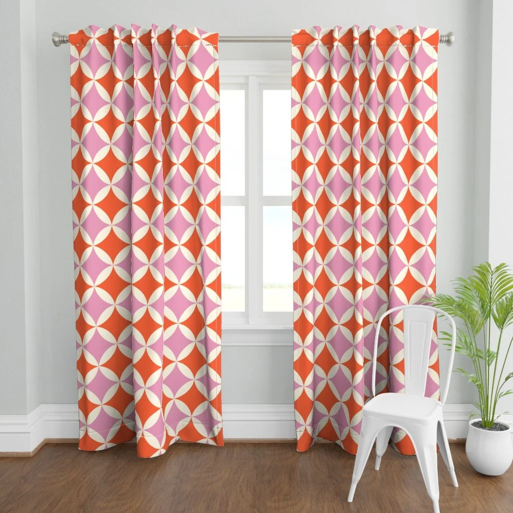 Curtains in a tangerine and pink orange peel quilt block print with cream petals, hung at a bright window, copyright Sandra Hutter.