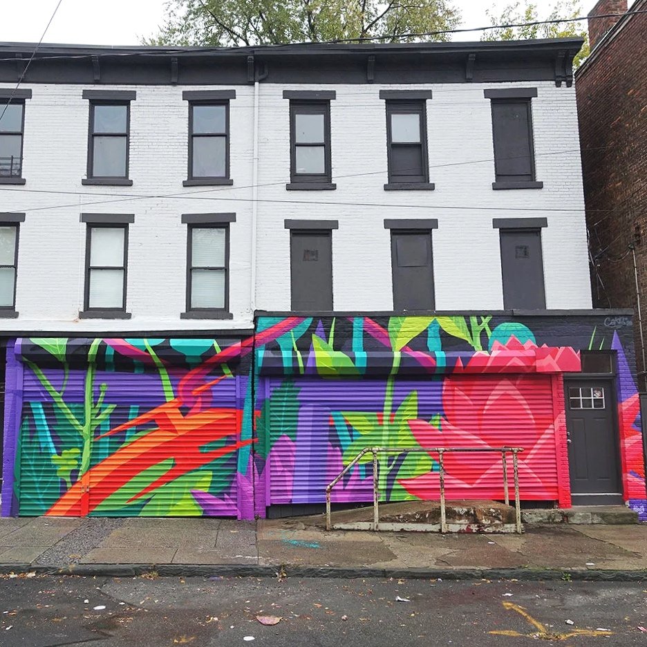 Colorful graffiti mural on a building's metal garage door, featuring abstract shapes and plant motifs in neon colors like green, pink, purple, and orange, with a black door and window above.