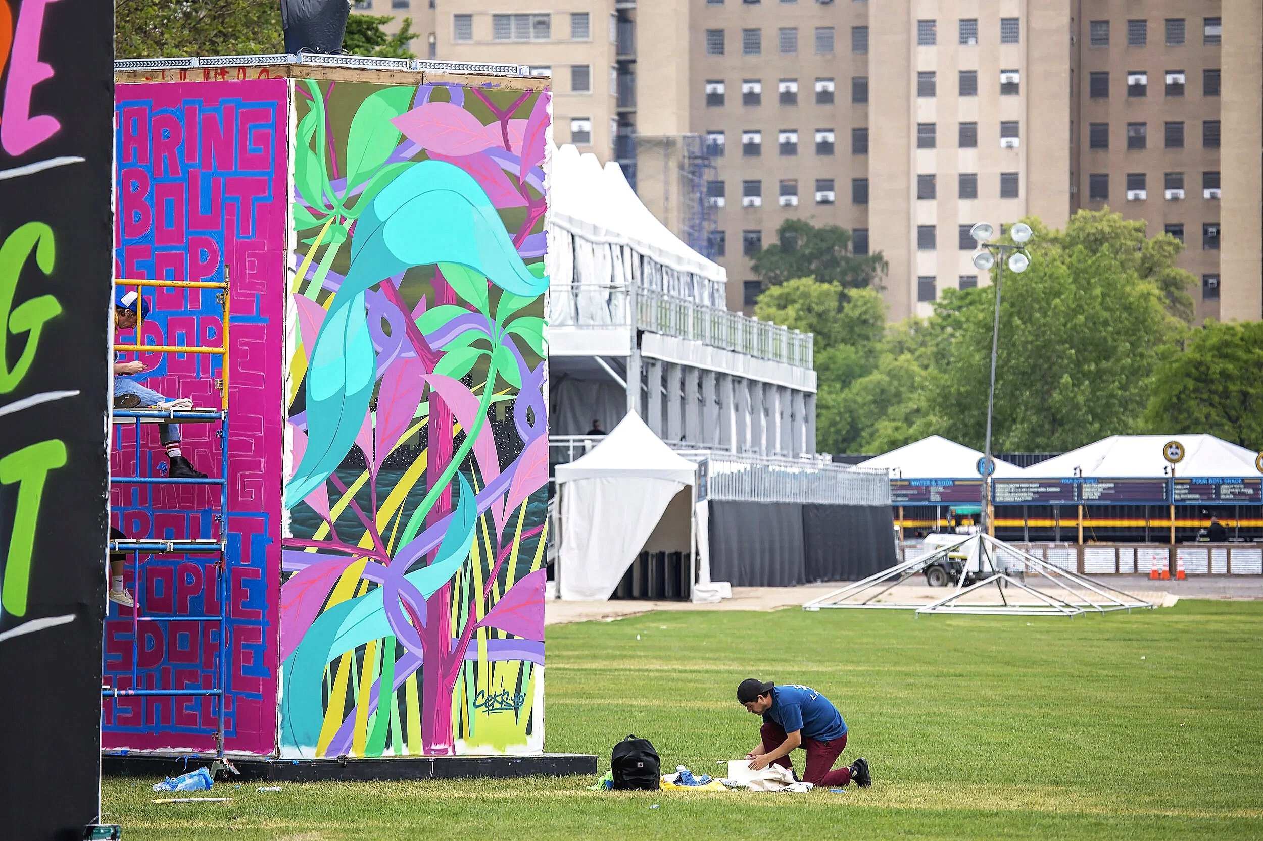 A man kneeling on the grass, working on art supplies near a large, colorful mural with tropical leaves. In the background, there are white tents, a tall building, and trees.