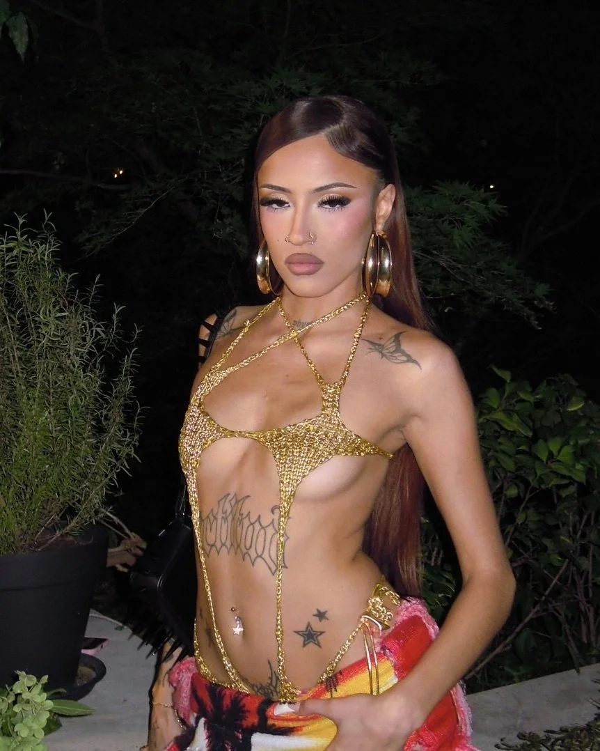 Mujer con cabello largo y suelto, vestido de gala con cadenas de oro, en un entorno oscuro con plantas, portando pendientes grandes y accesorios visibles.