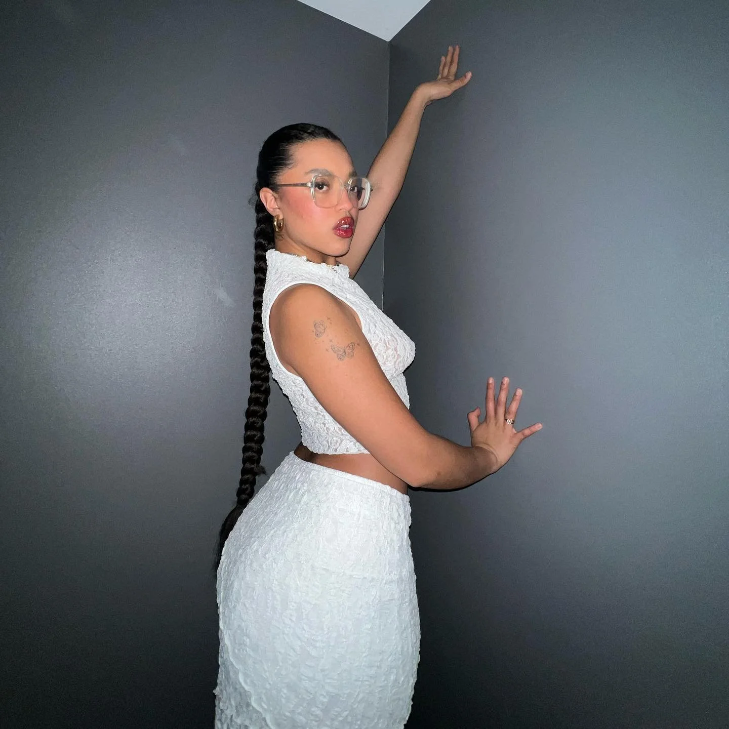 Mujer con cabello largo trenzado, vestido blanco de encaje, gafas transparentes, y tatuajes en el brazo, posando contra una pared gris con una mano en el muro y la otra levantada.