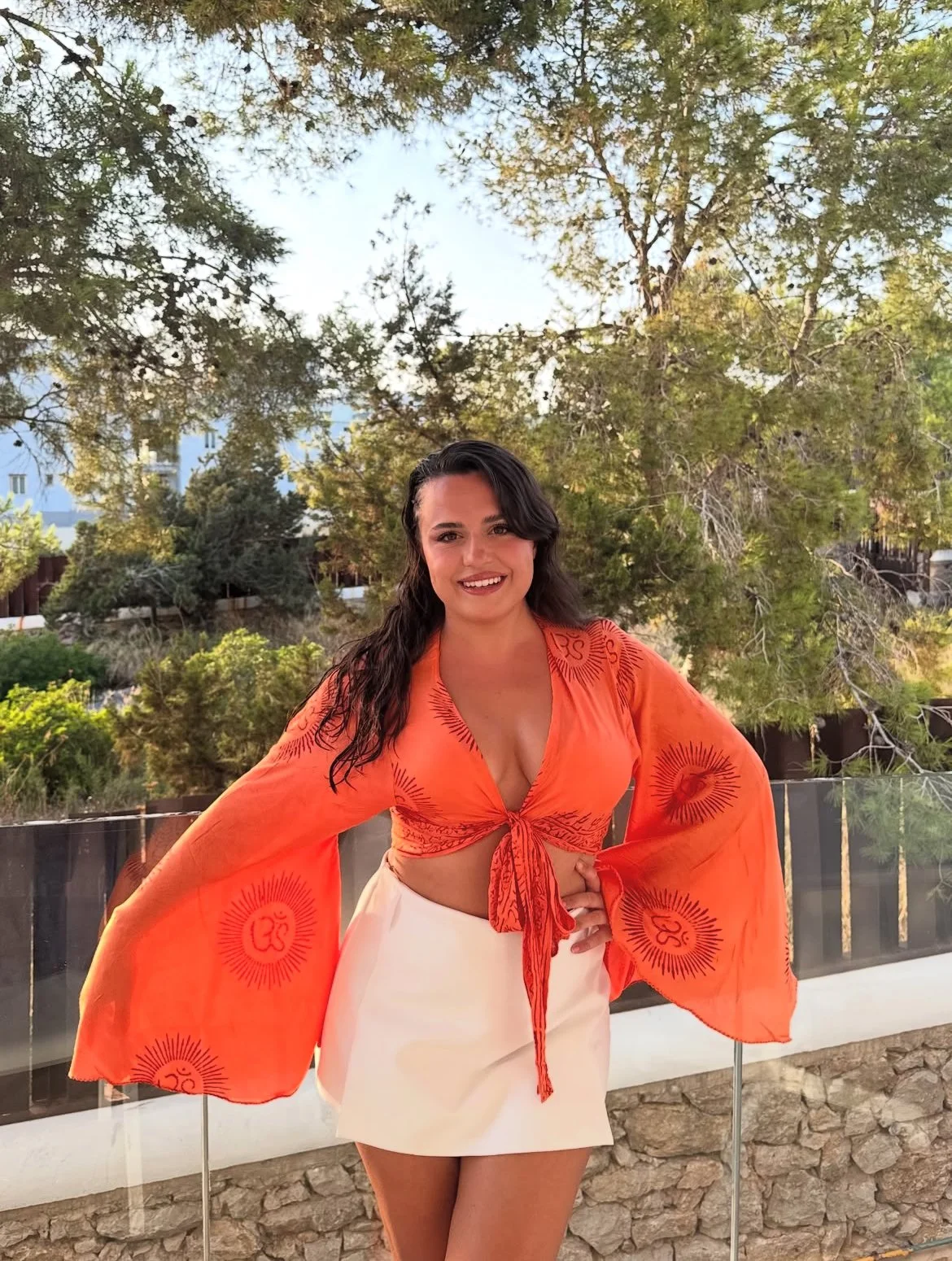 Una mujer con cabello castaño oscuro y sonrisa, usa un top naranja con estampado y una falda blanca, de pie en un balcón con fondo de árboles y cielo.