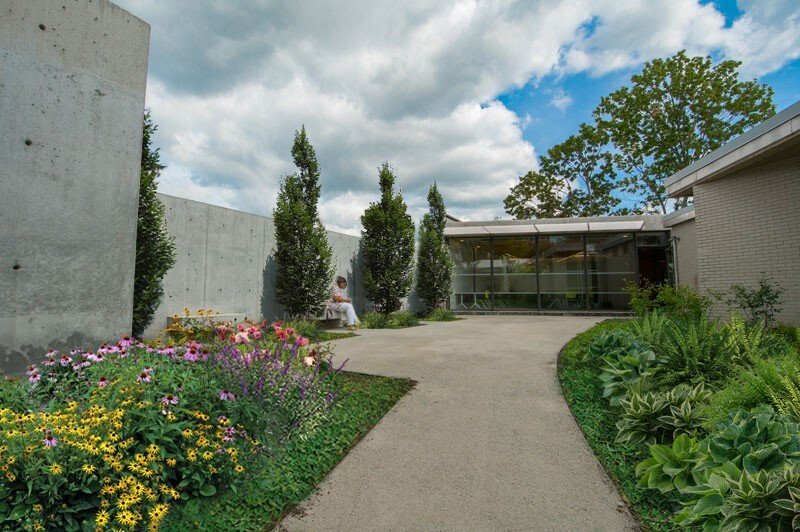 A courtyard garden with colorful flowers, tall trees, and modern buildings, featuring a person sitting on a bench.