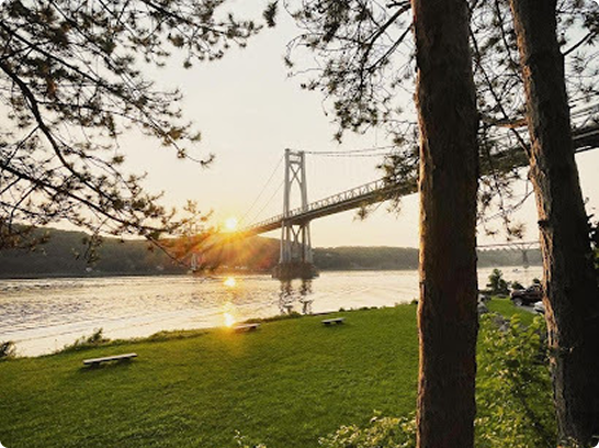Sunset over a river with a bridge, trees in the foreground, and a grassy park area.