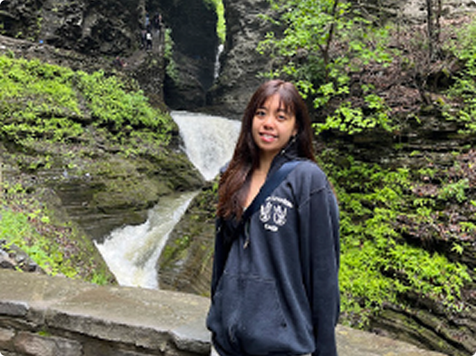 Young woman standing in front of a waterfall with lush greenery in a rocky gorge.