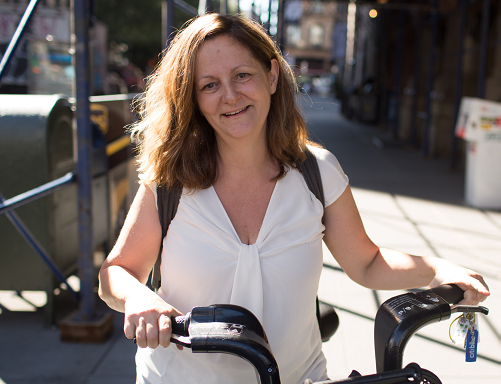 Smiling woman in a white shirt riding a bicycle outdoors in a city street