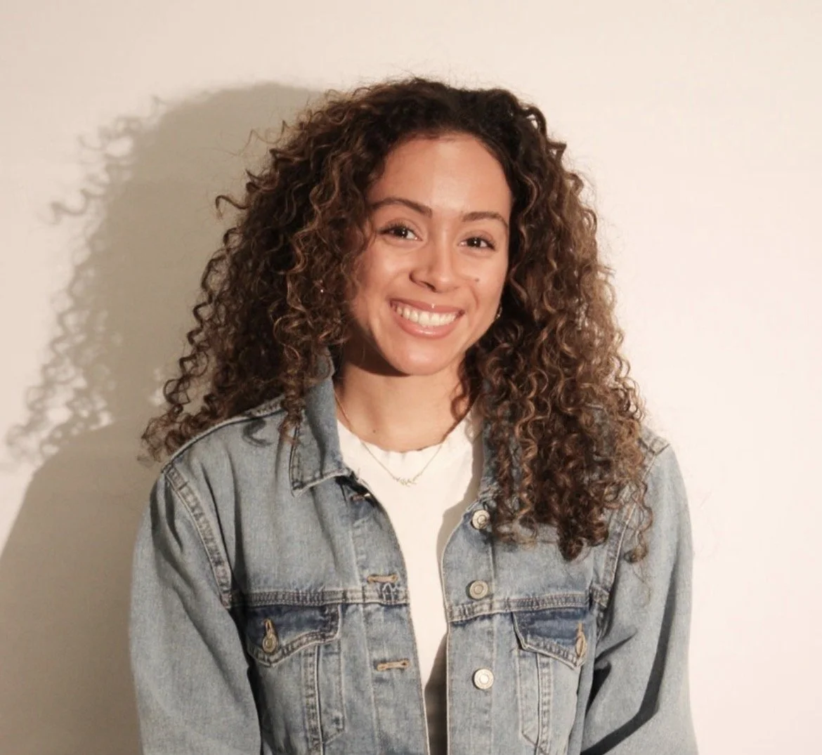 A person wearing a blue jacket and white t-shirt with long, curly hair smiles in front of a beige background