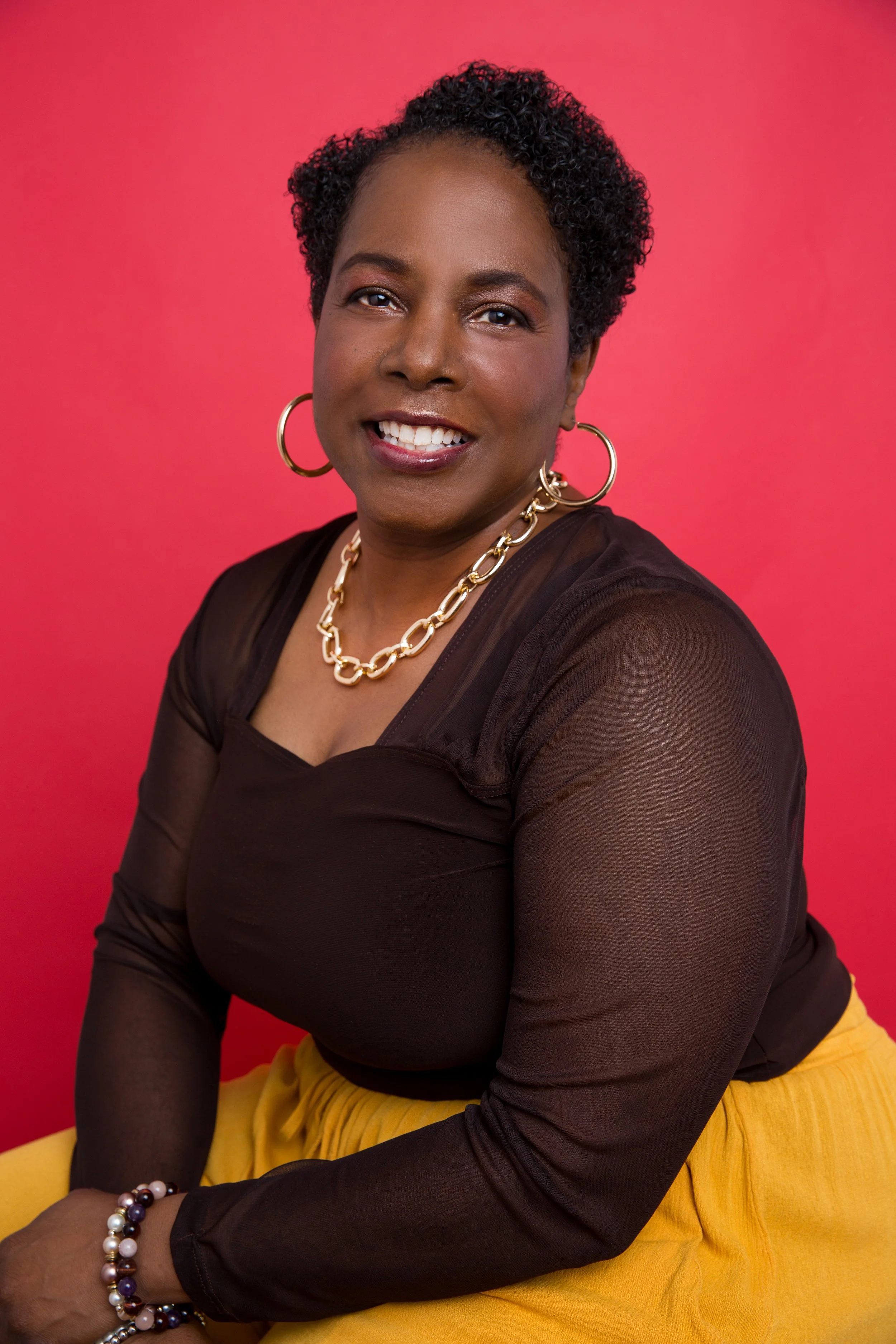 A person with short hair wearing a black shirt, a gold chain necklace and hoop earrings smiling in front of a red background