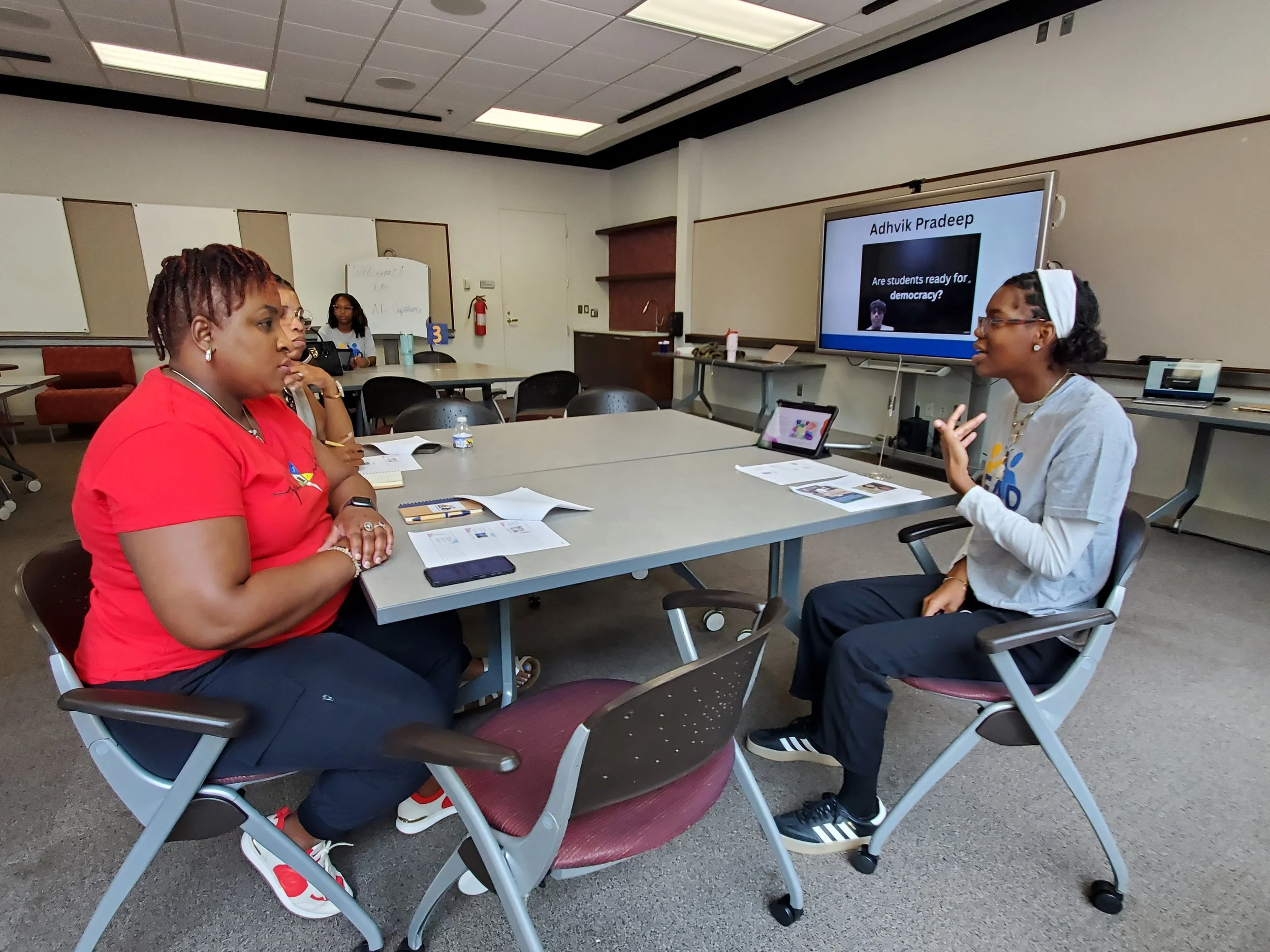 A student in a grey shirt sitting at a table presenting to someone else at the table who is wearing a red shirt