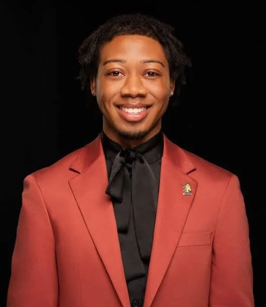 A person wearing a read suit jacket and a black shirt smiling in front of a dark background