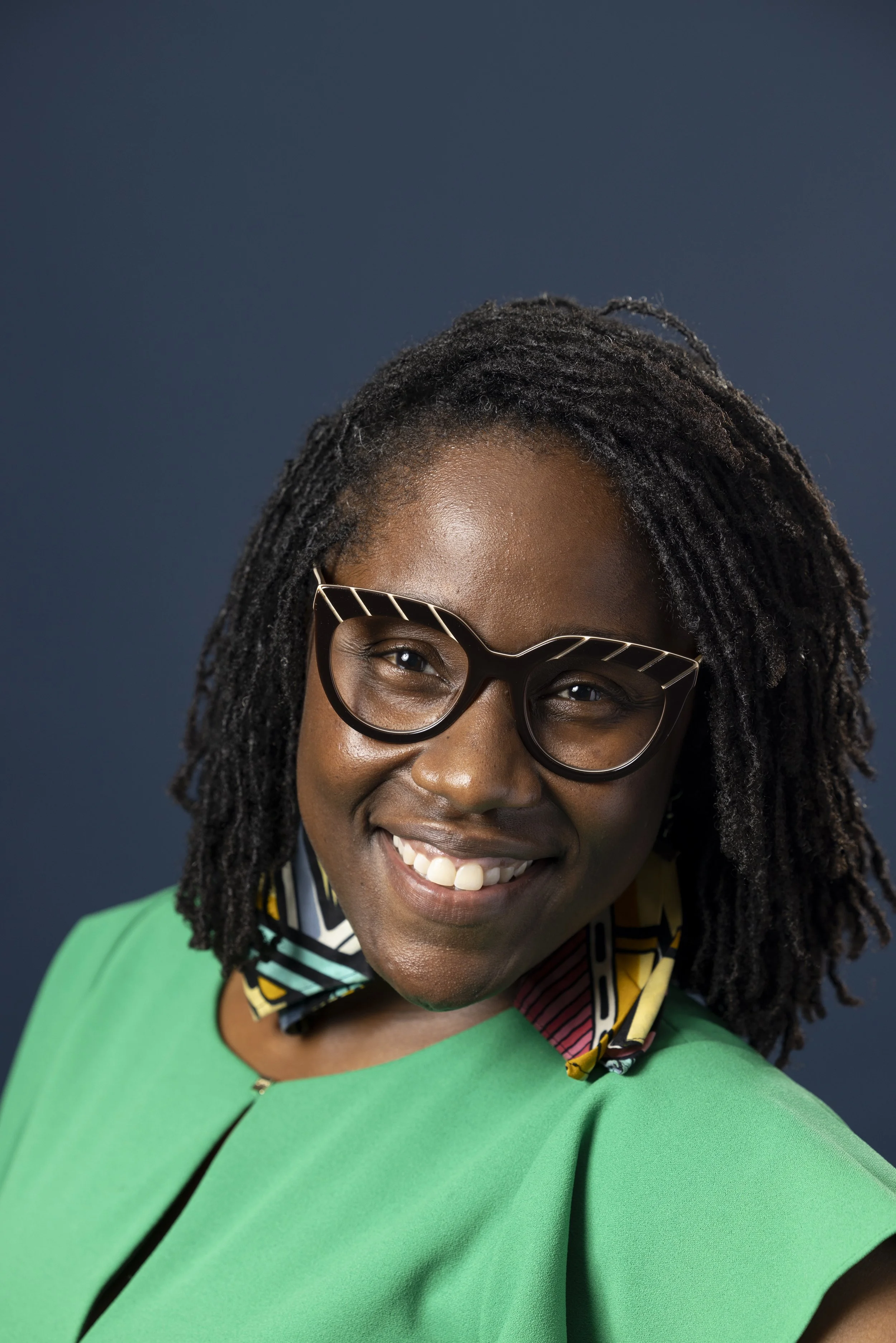 A person wearing dark glasses and a green shirt smiling in front of a dark blue background
