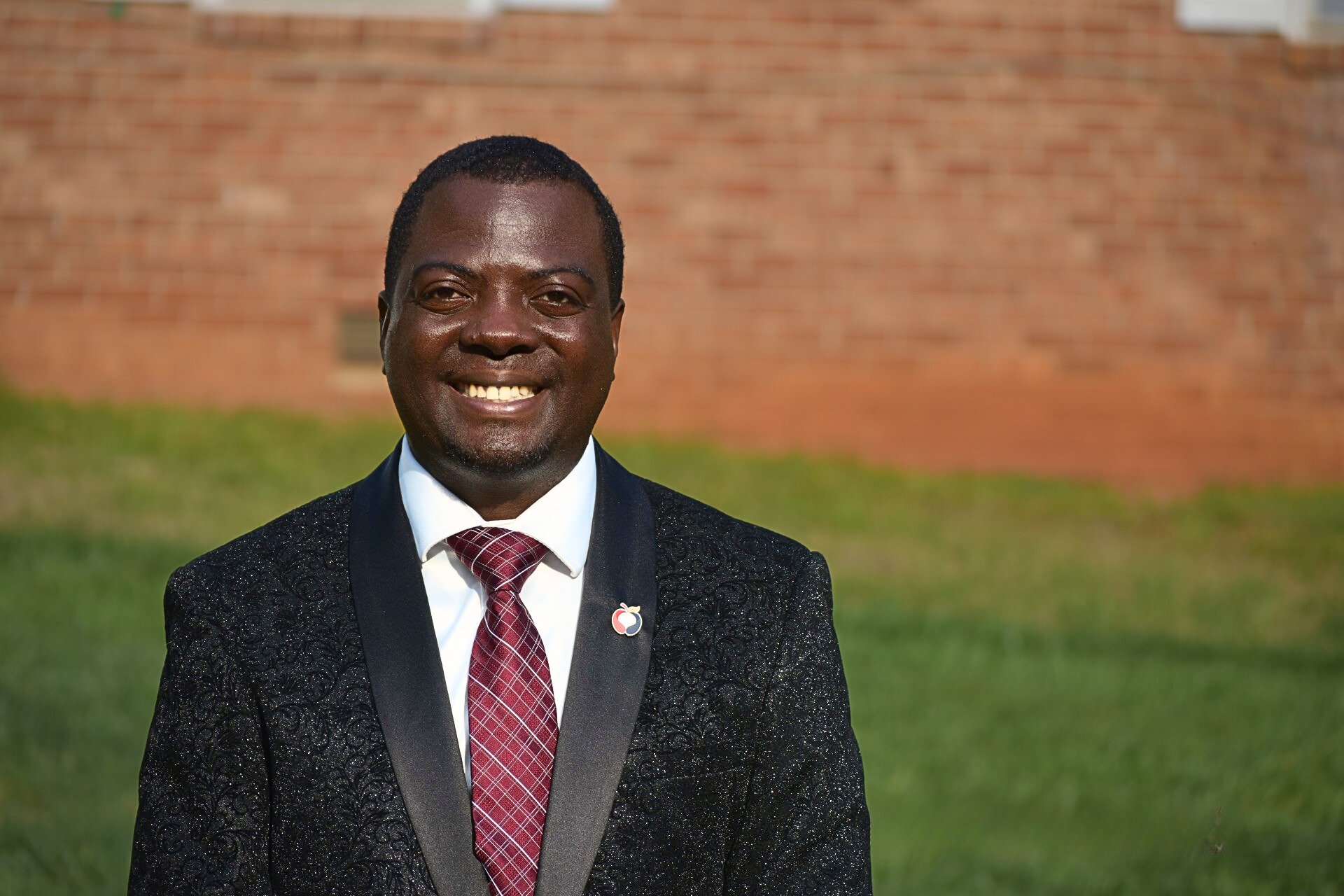A person wearing a dark suit and a red tie smiling in front of a brick wall background
