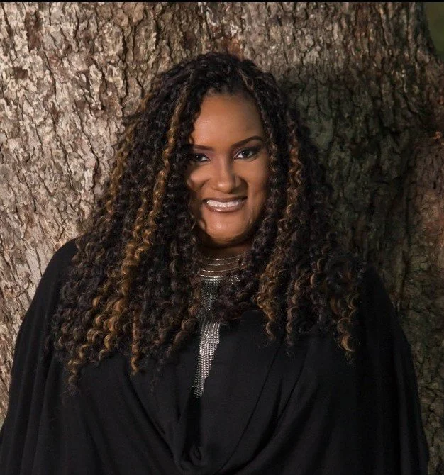A person wearing a black shirt with long dark hair smiling in front of a wood background