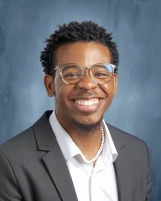 A person wearing glasses, a dark blazer, white shirt, and a beaded necklace smiles in front of a blue-gray background.