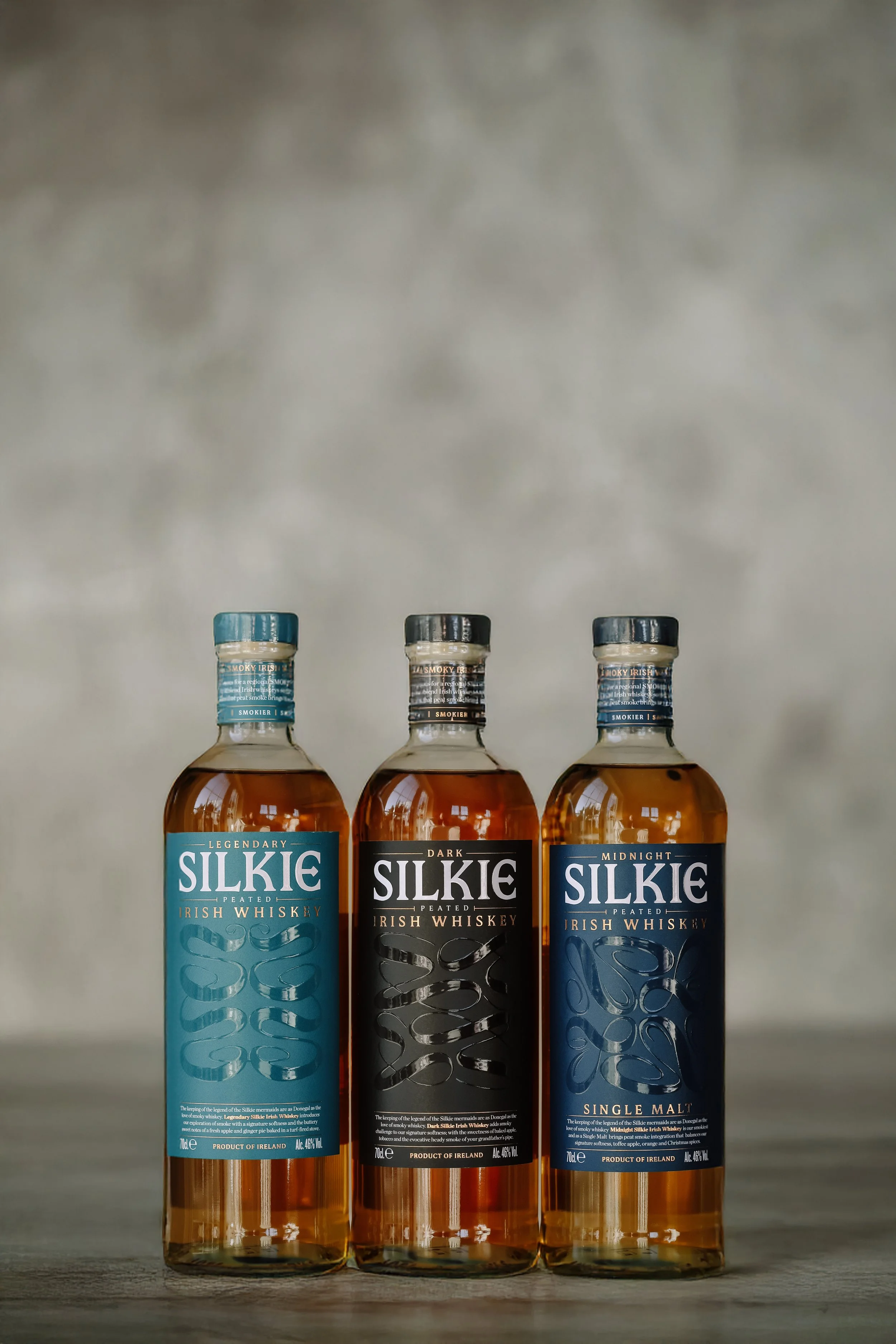 Three bottles of Silkie Irish Whiskey on a wooden surface with a blurred gray background.