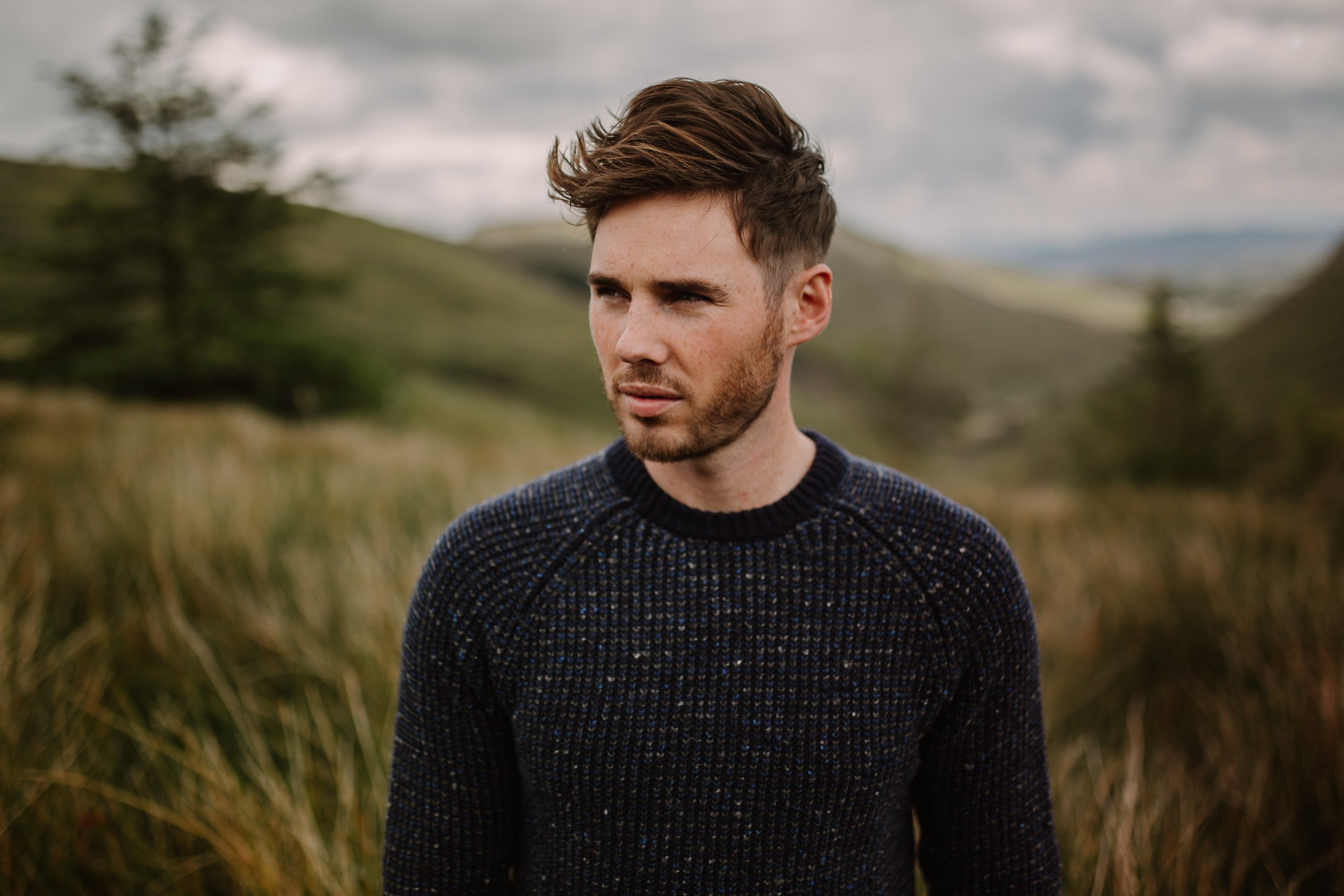 Young man with brown hair and beard standing outdoors in a field with tall grass and hills in the background.