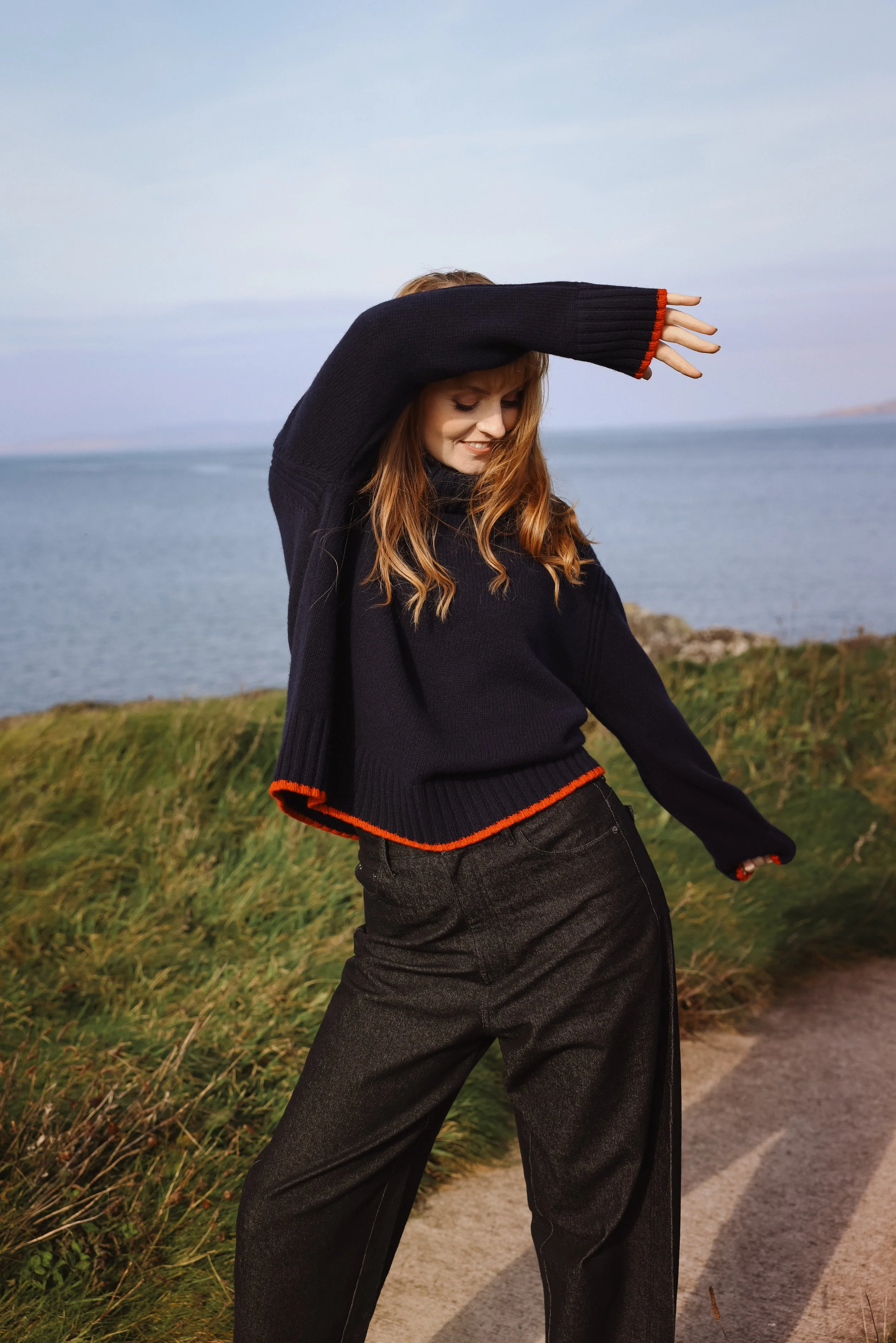 A young woman with red hair, smiling, standing outdoors by a body of water with grassy dunes in the background. She is wearing a black sweater with red trim and black pants.