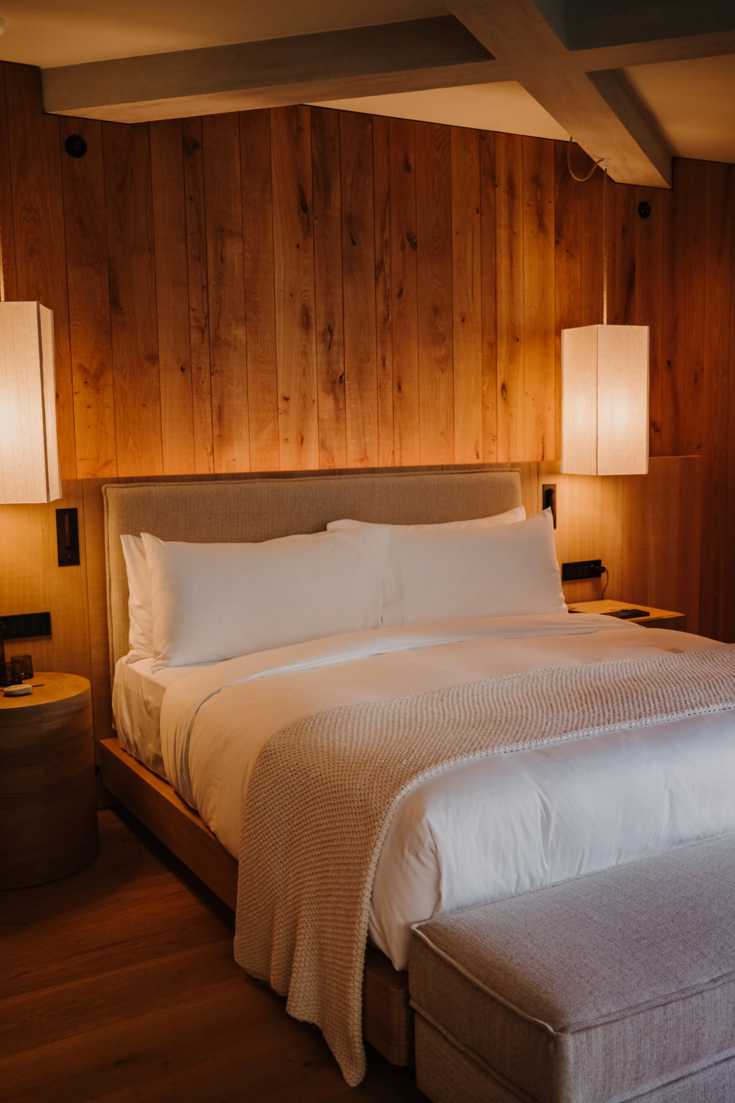 A bedroom with a large bed surrounded by wood-paneled walls, illuminated by two hanging bedside lamps.