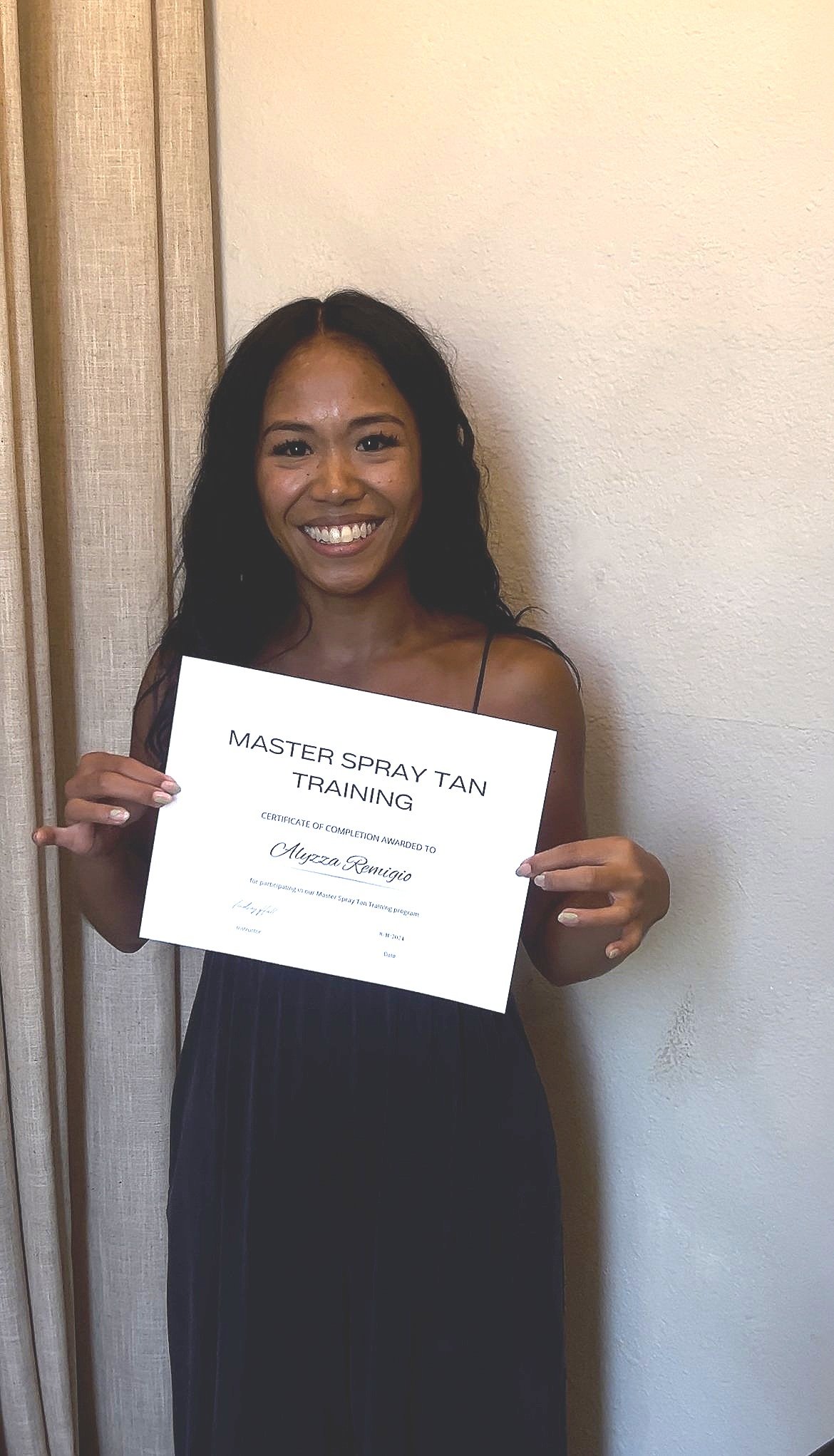 Woman smiling and holding a certificate that reads 'Master Spray Tan Training'. She is standing in front of a beige wall and dressed in a black sleeveless dress.