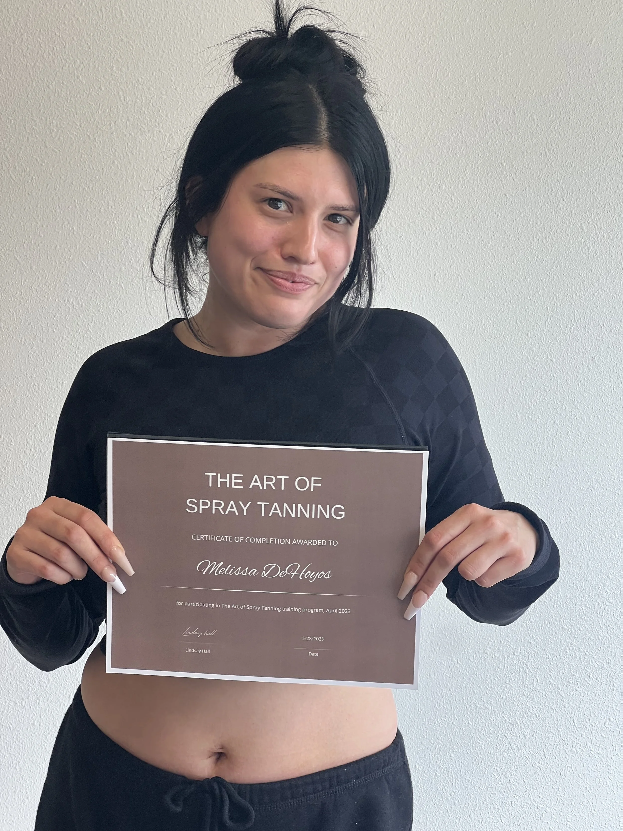 A woman with black hair styled in a messy bun holding a certificate that reads 'The Art of Spray Tanning' and 'Melissa DeHoyos', smiling lightly against a plain white wall.