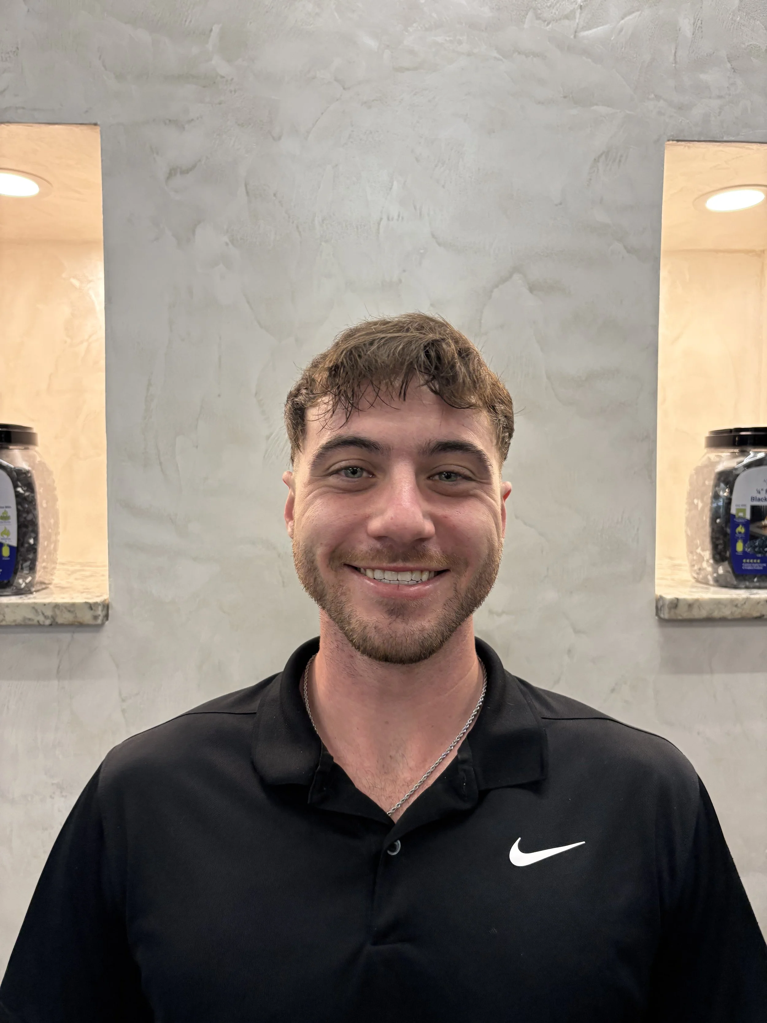 A smiling man with short brown hair, light facial hair, wearing a black Nike polo shirt and a silver chain, standing in front of a textured gray wall with two illuminated niches containing jars of colorful candy.
