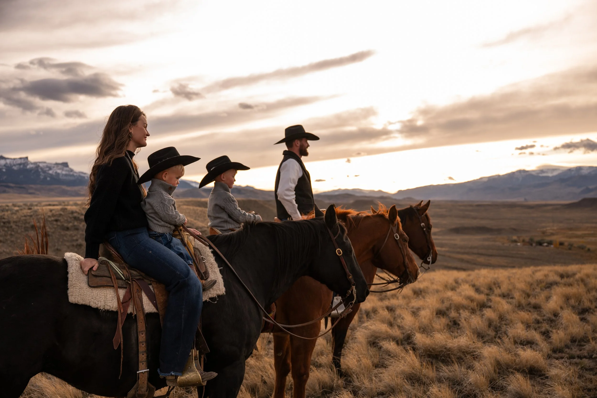 Family of five riding horses across open plains at sunset, mountain range in background, with a woman and children wearing cowboy hats.