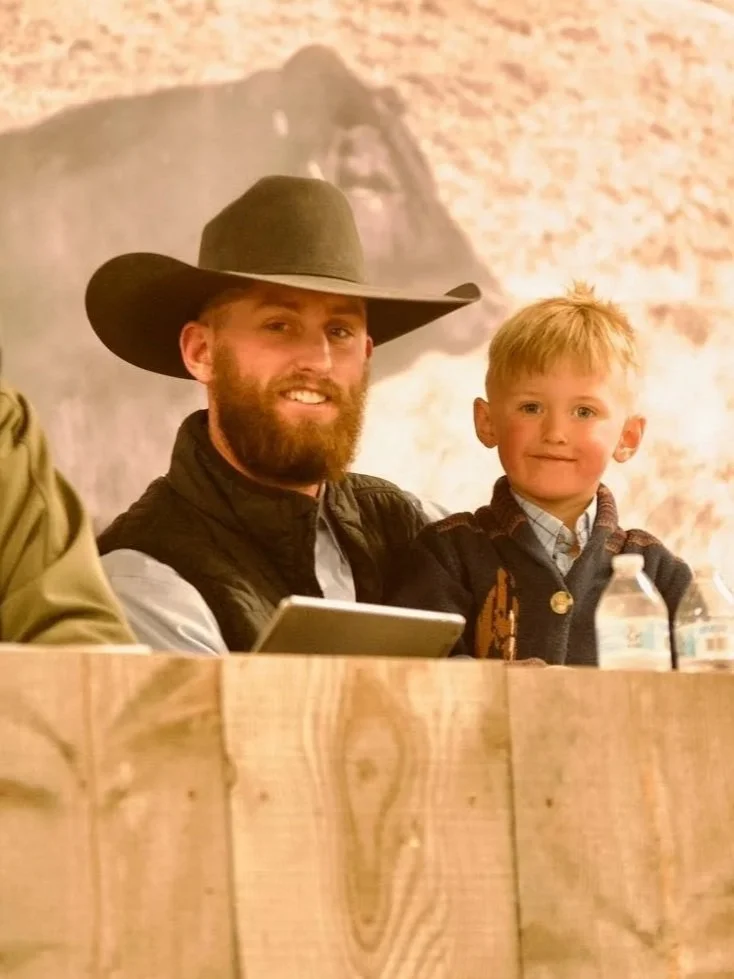 A man with a beard wearing a cowboy hat and a young boy sitting at a table with water bottles, both smiling.