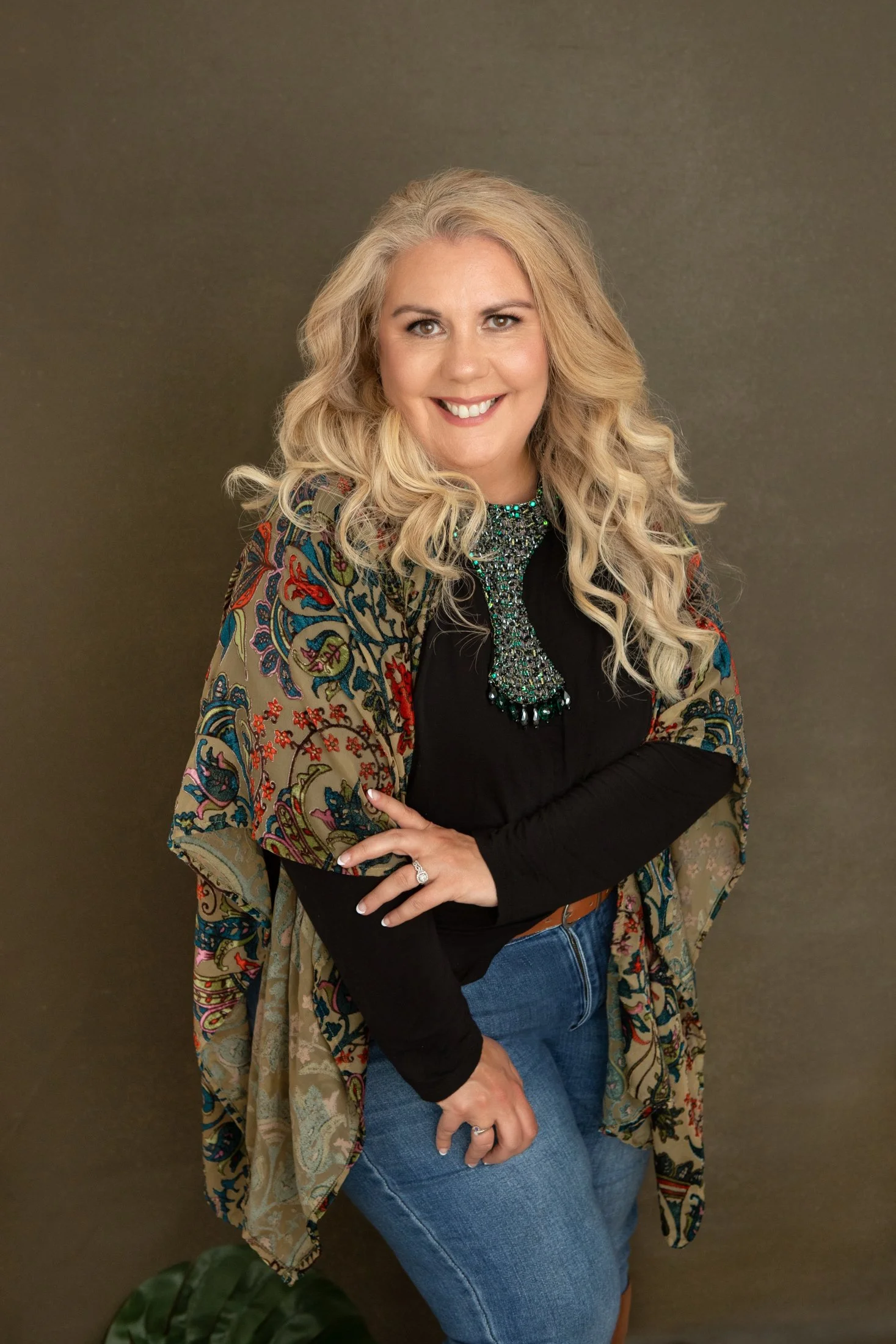 owner of gold hummingbird LLC. Blonde woman, brown eyes, long black shirt, floral patterned shawl, jeans.