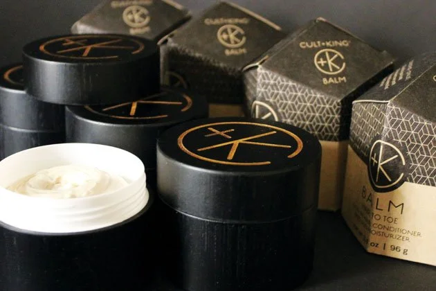Several black containers of CultLUX Balm, a hair or skin product, one open with white cream inside, alongside multiple boxed packages of the same product.