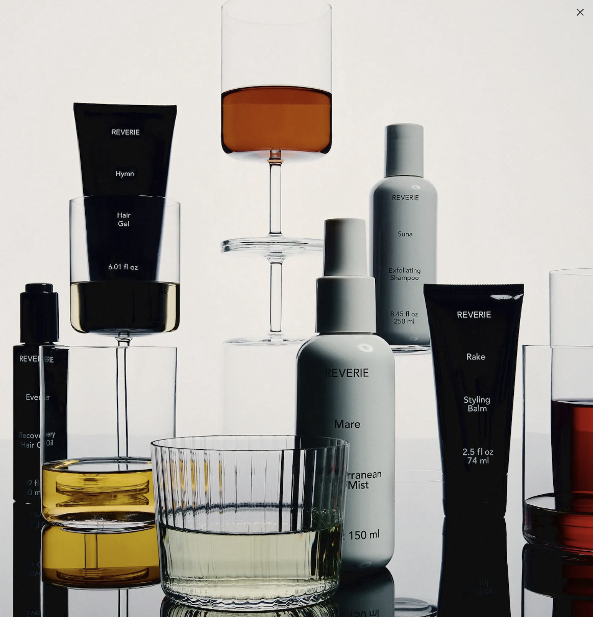 Arrangement of skincare and hair care products including bottles, tubes, and glasses, some with liquids in shades of yellow, amber, and red, displayed on transparent and glass surfaces.