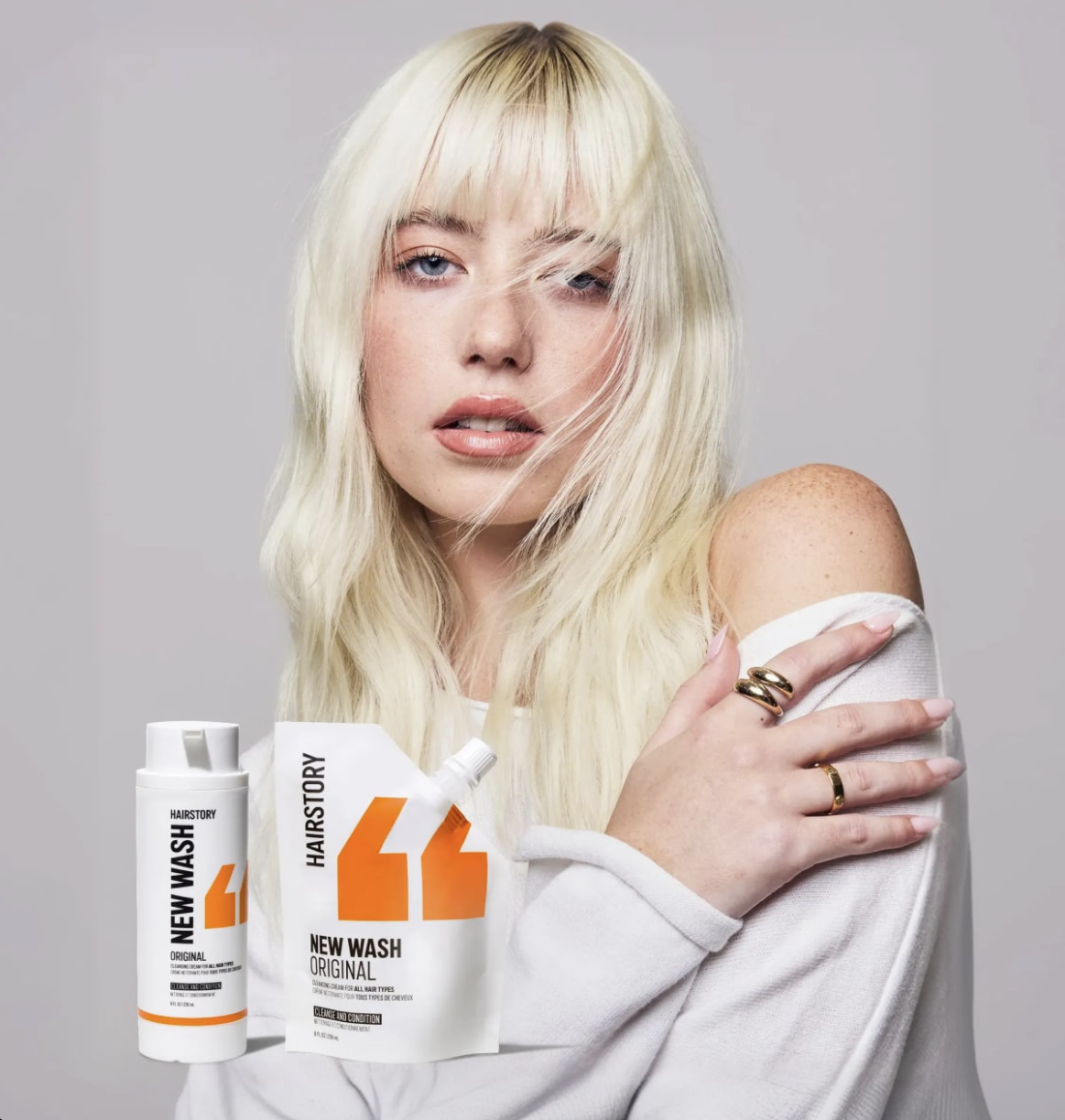 A woman with long blonde hair and fair skin holds her left shoulder with her right hand. She wears a white off-the-shoulder top with gold rings and has a confident facial expression. Next to her are hair care products labeled "New Wash Original" by Hairstory.