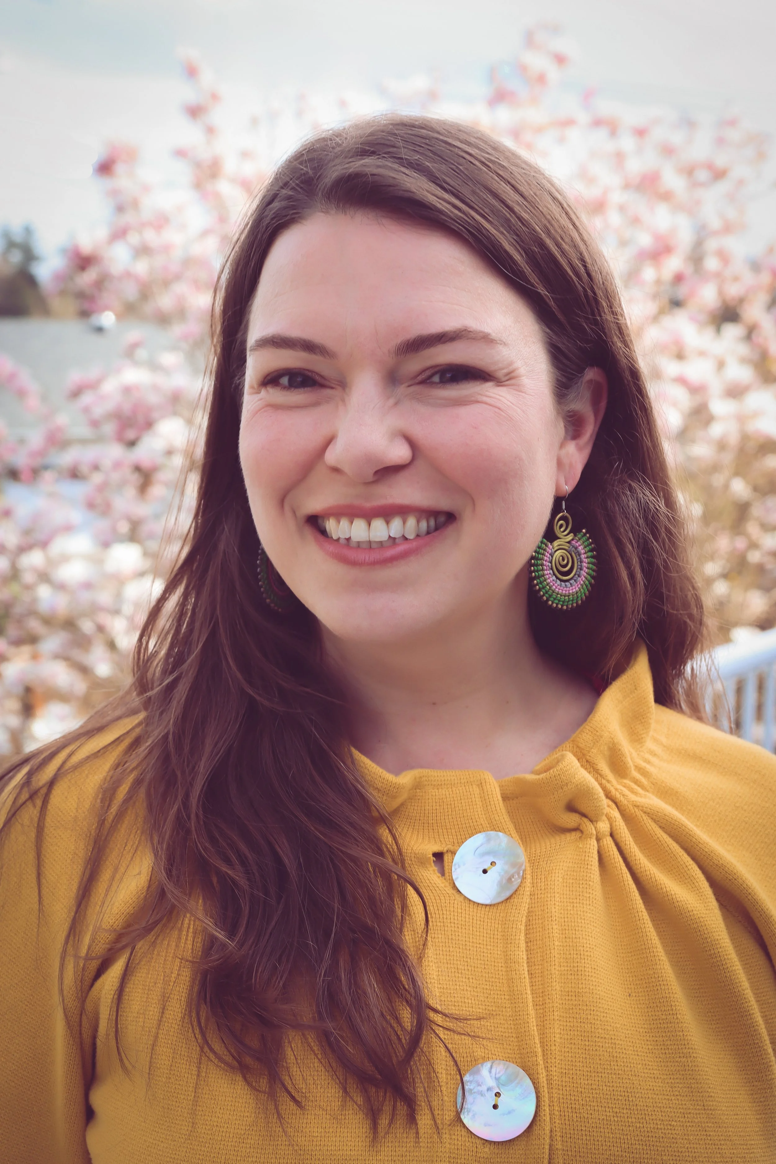 Headshot photo of Leah, standing outside in front of a pink flowering tree and wearing a yellow top