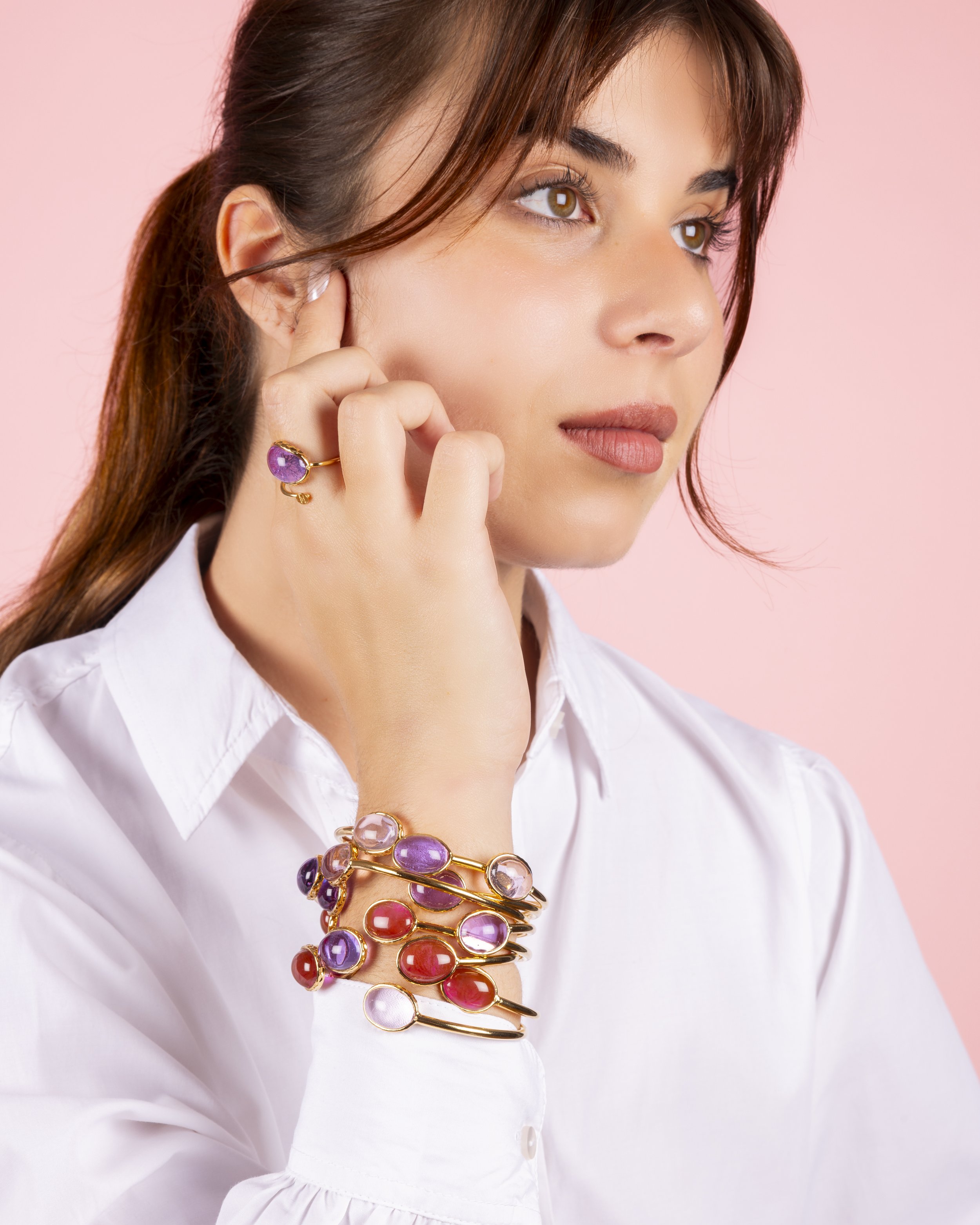 Woman wearing bracelets and a ring with colored stones, pink background.