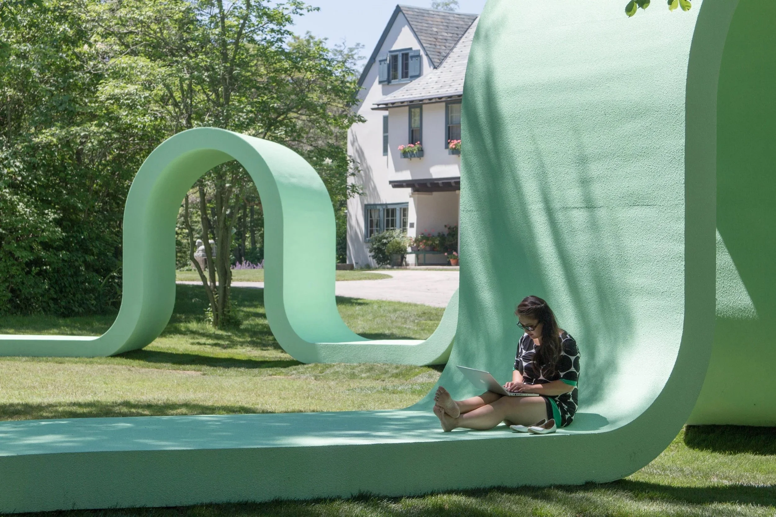 A woman sits on a massive, winding light-green ribbon sculpture in a grassy yard, using her laptop in front of a traditional house.