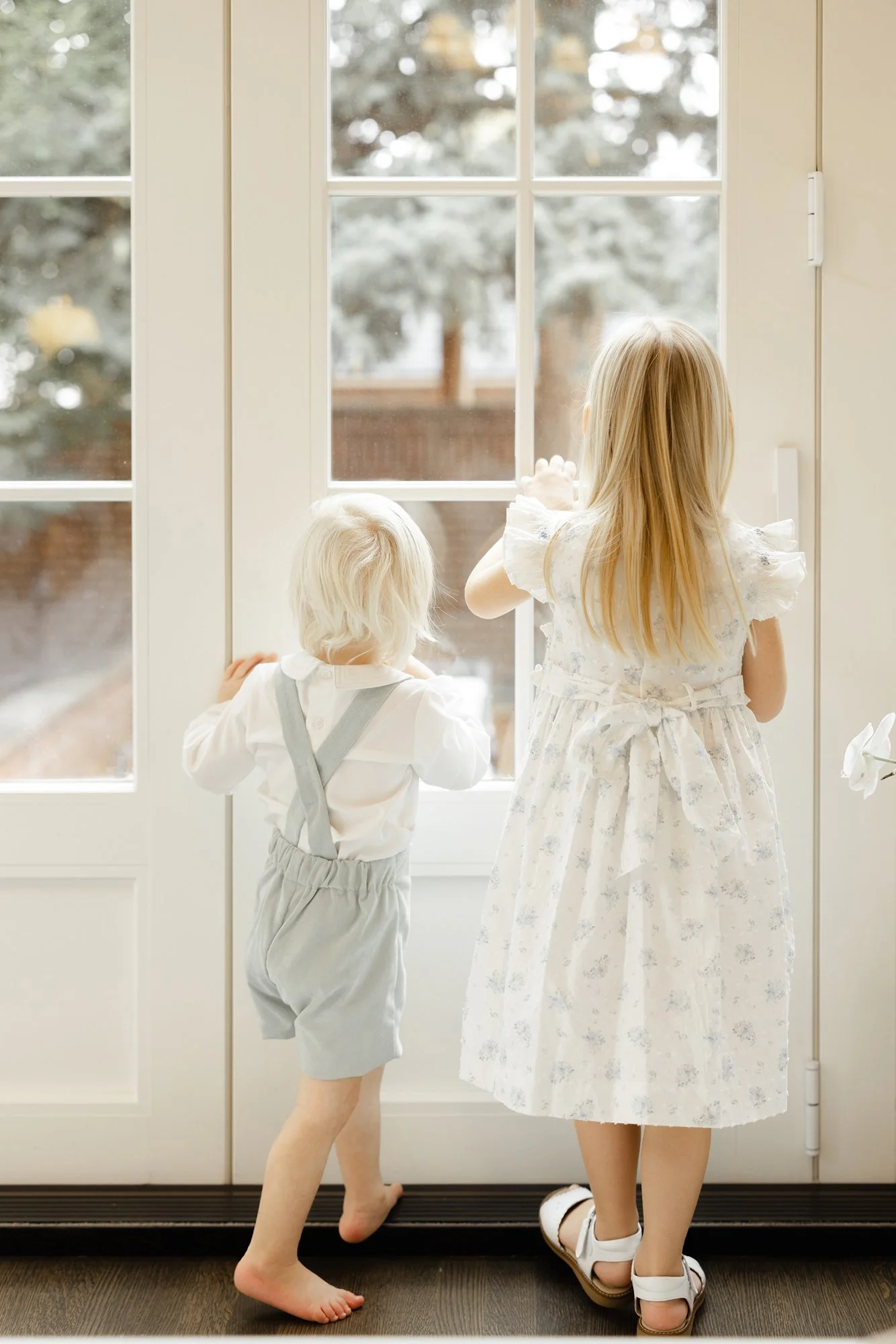 Two young siblings looking out a bright window during their family's newborn session.