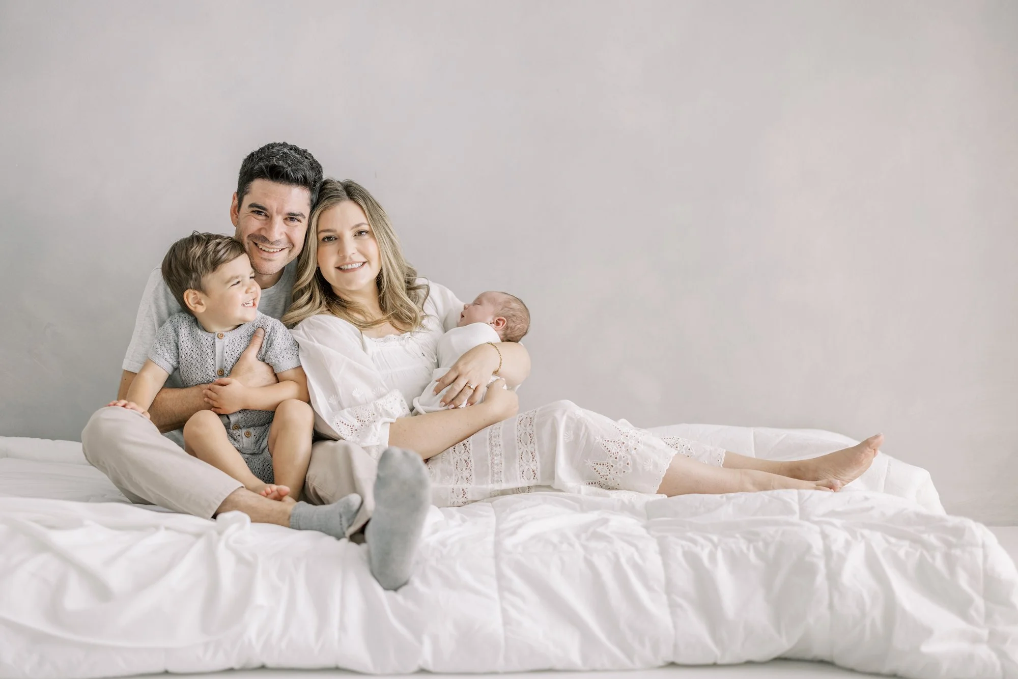 Parents and toddler cuddling together on a bed with their newborn during a relaxed studio family portrait.