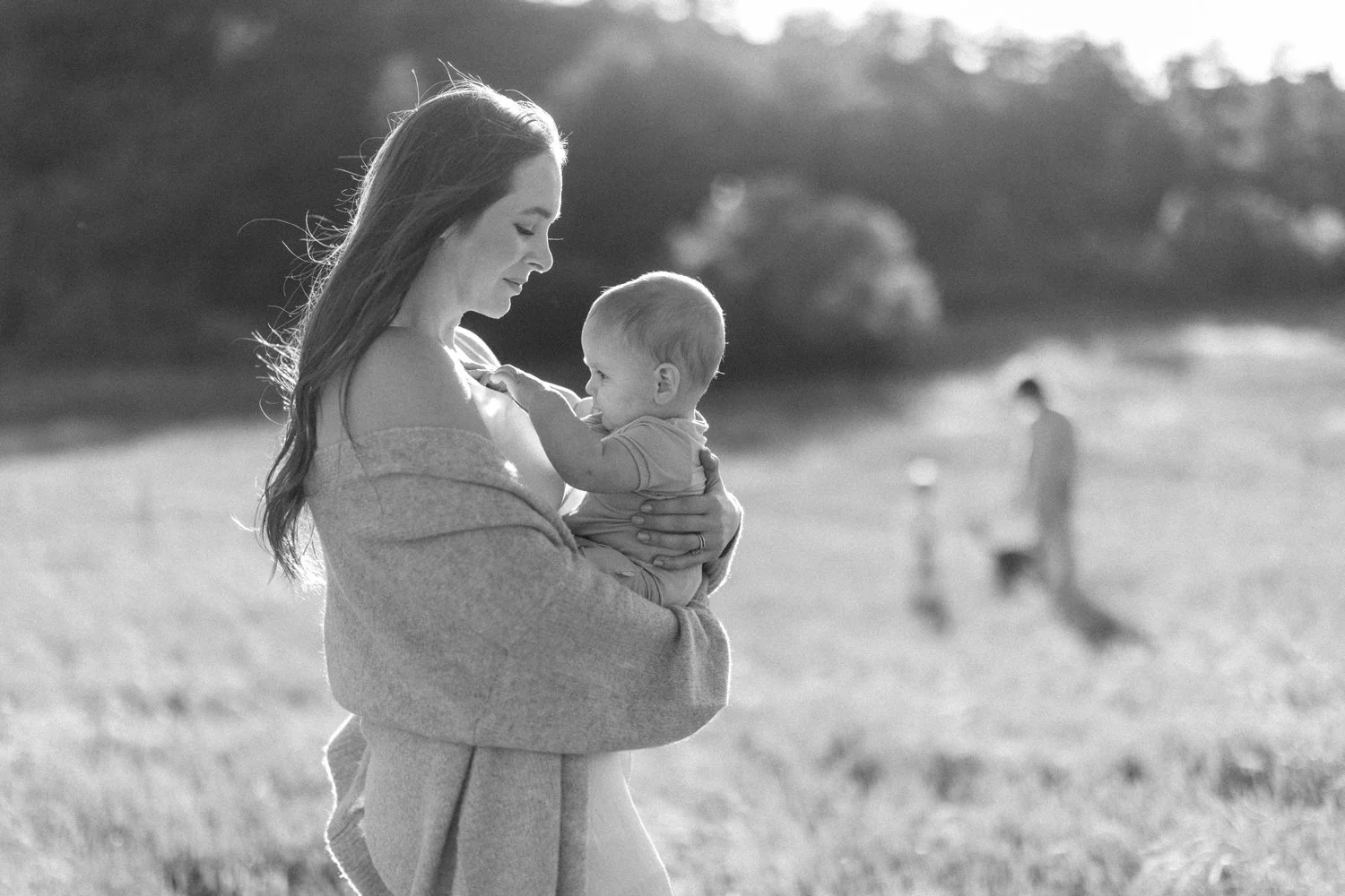 Black-and-white portrait of a mother holding her young child with children playing in the field behind them.