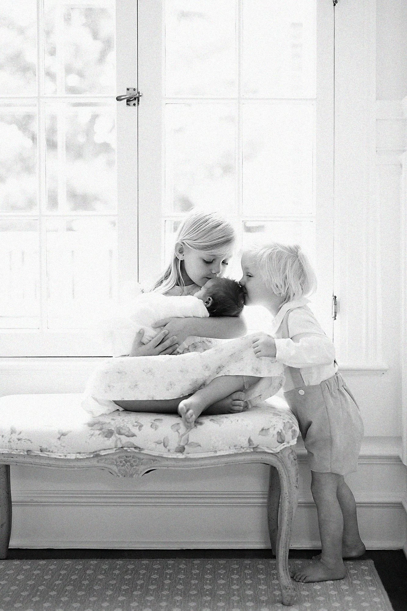 Older brother and sister sharing a gentle kiss with their newborn sibling during a natural light family newborn session.